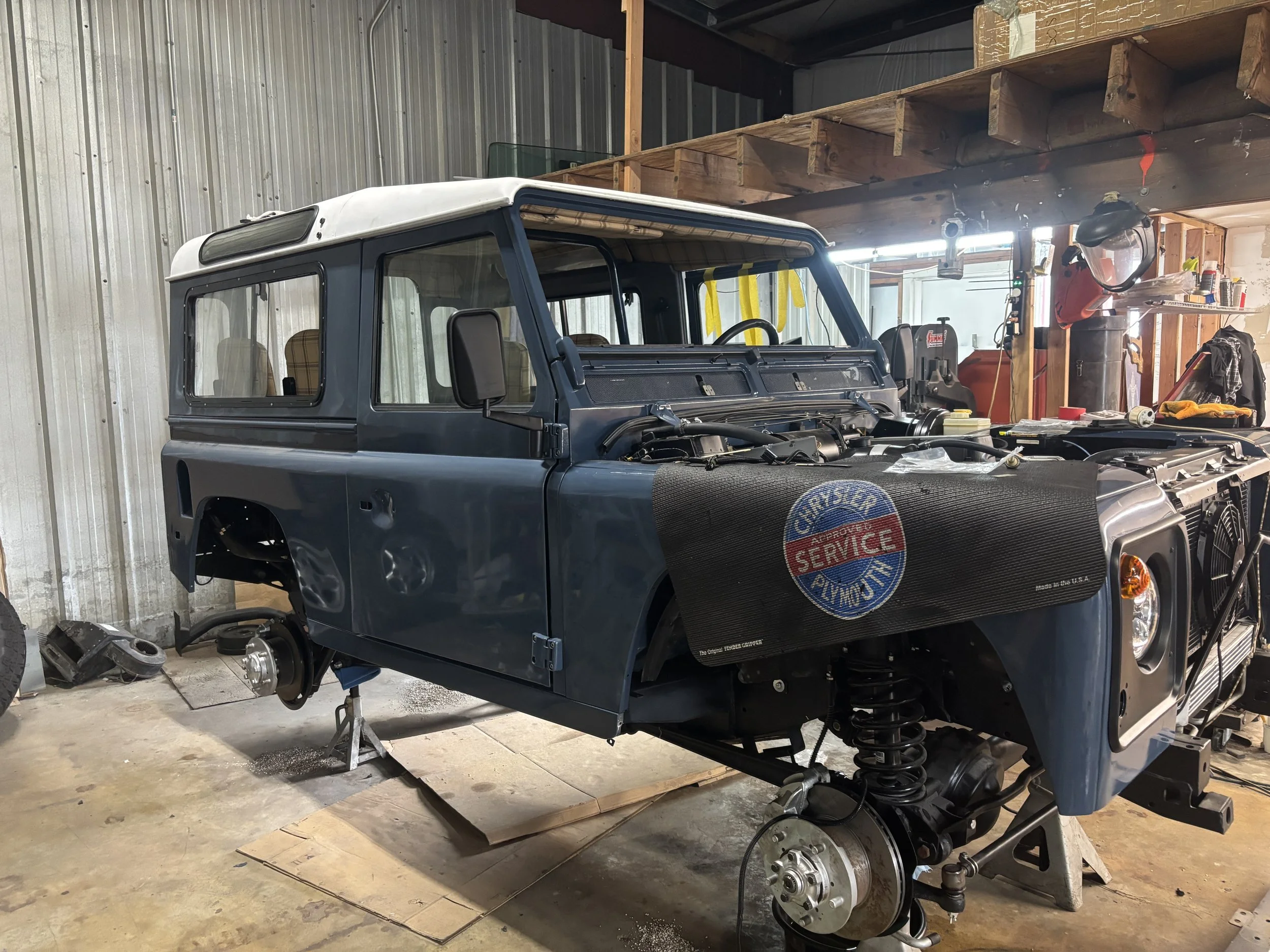 The front of a vintage Jeep vehicle in a garage, with no wheels and part of its engine visible. The garage has wooden shelves and various tools and supplies along the walls.
