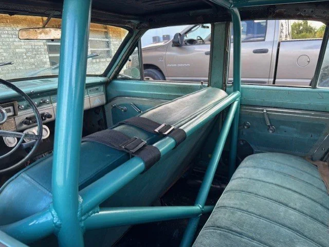 Interior of a vintage car with a roll cage, a padded bench seat, and a large metal bar running across the front.