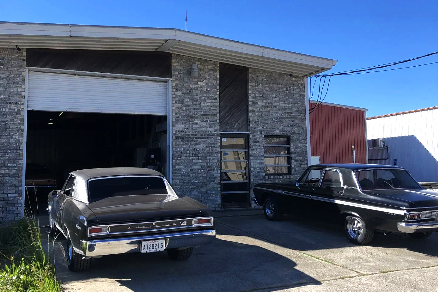 Two vintage black cars, 1966 Chevrolet Chevelle, and a 1965 Plymouth Fury, parked outside a brick building with an open garage door on a sunny day.