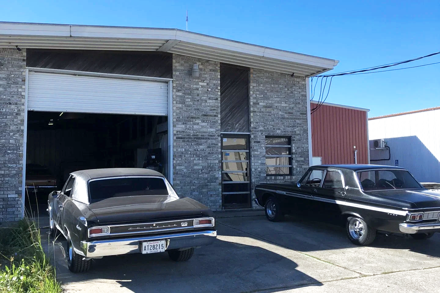 Two vintage cars parked outside a brick building with a large open garage door on a sunny day.