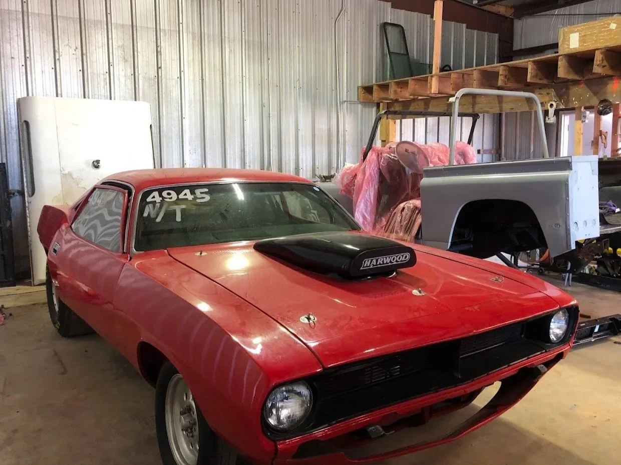 A red drag racing car with a black hood scoop, parked in a garage or workshop with automotive parts and tools around.