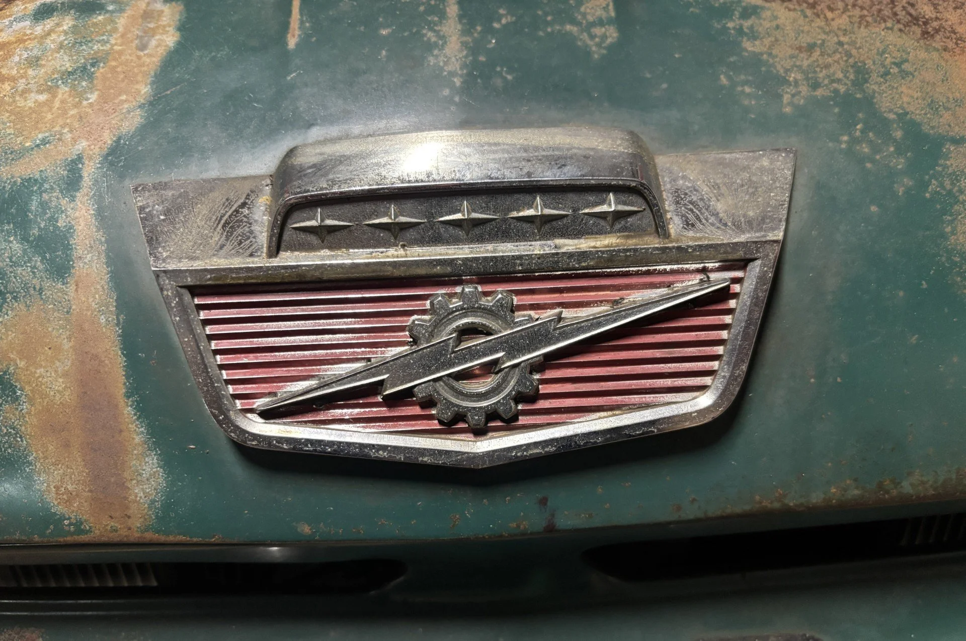 Close-up of a vintage fire truck emblem with a chrome badge featuring a gear and lightning bolt, mounted on a worn, rusty green surface.