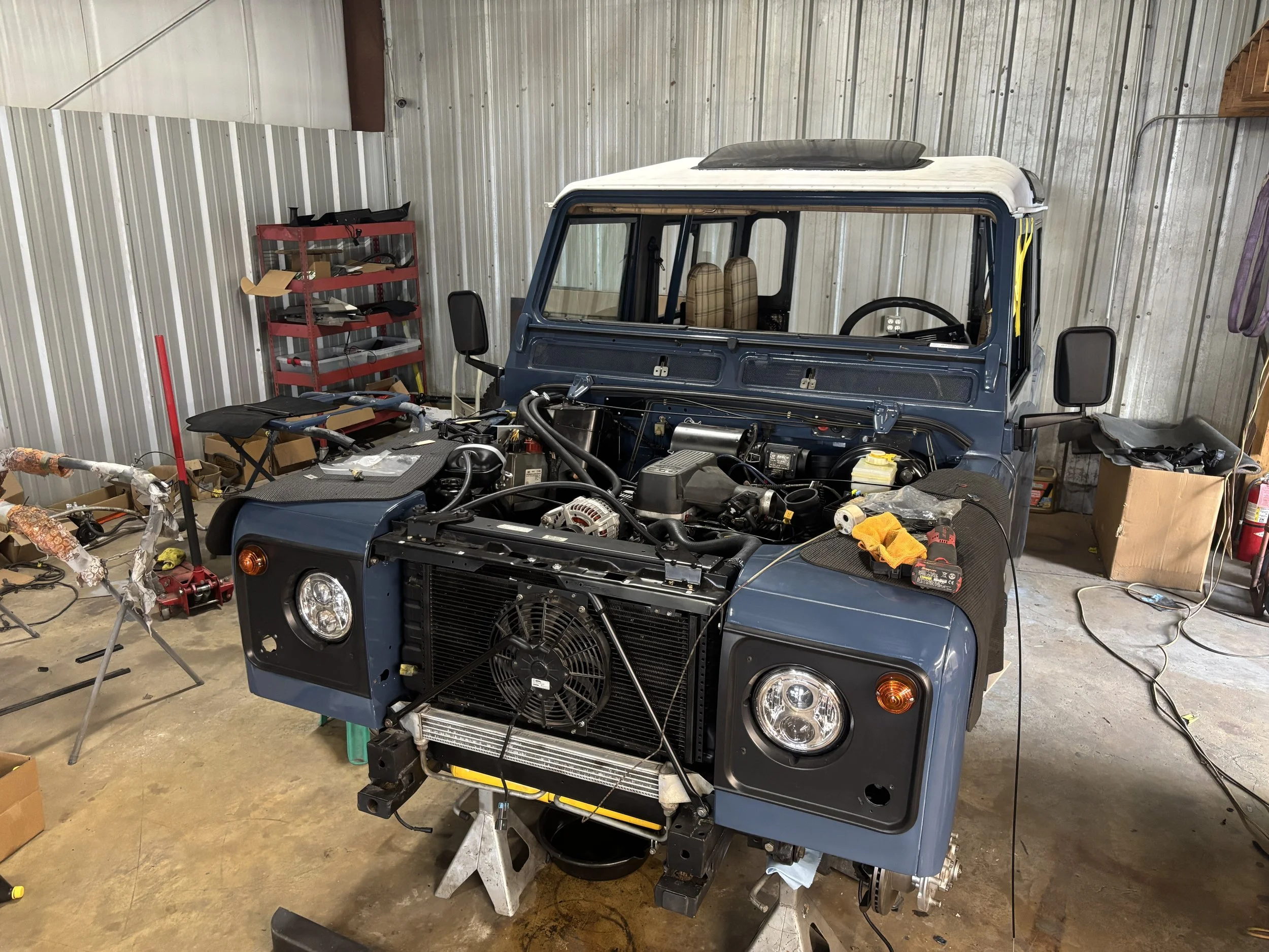 A blue off-road vehicle in a garage with its front panel removed, exposing the engine. The garage has a metal wall, a red shelf with various tools and parts, and clutter including boxes, tools, and a yellow cloth.