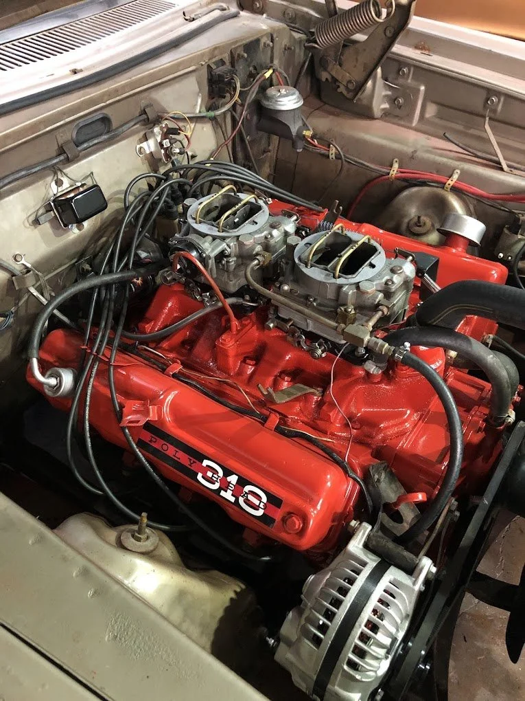 A close-up view of a car engine with a bright red valve cover that says 'Toyota 318'. The engine has exposed components and wiring, with an alternator visible at the bottom right and various mechanical parts surrounding it.