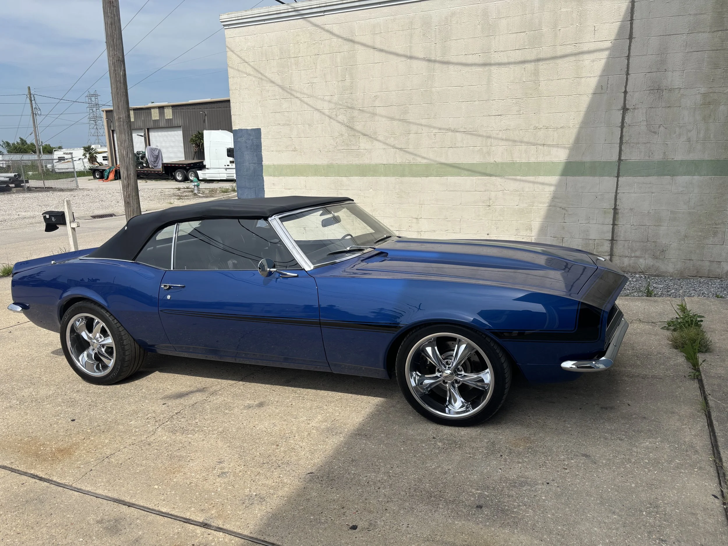 A blue vintage convertible car parked outside next to a beige brick wall with a green stripe. The car has a black soft top and shiny chrome wheels.