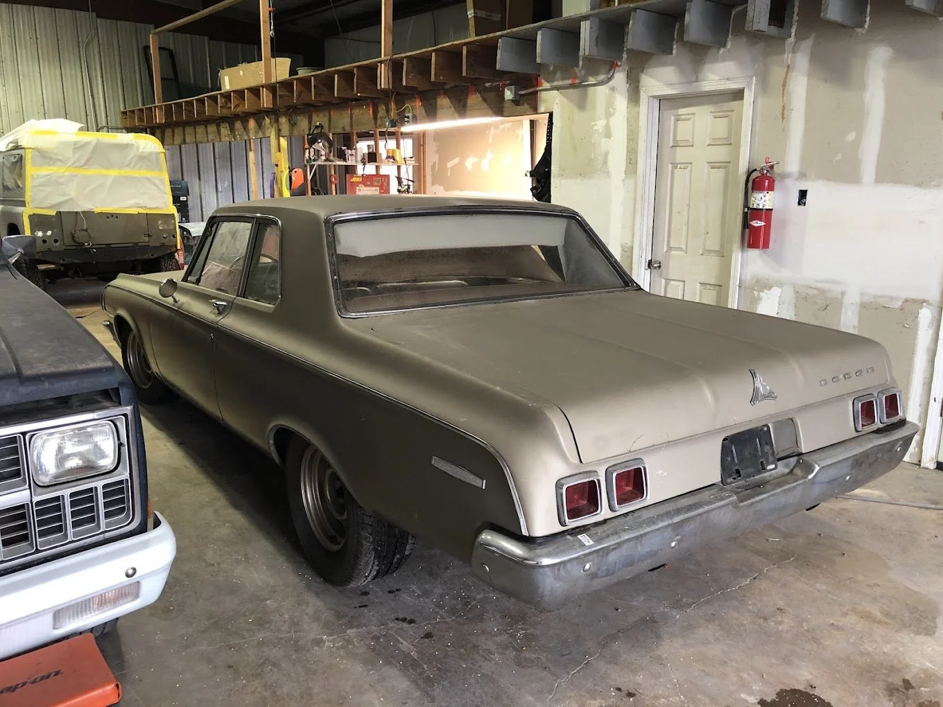 A vintage beige Dodge car in a garage, surrounded by other vehicles, with a fire extinguisher on the wall, and woodworking tools and equipment in the background.