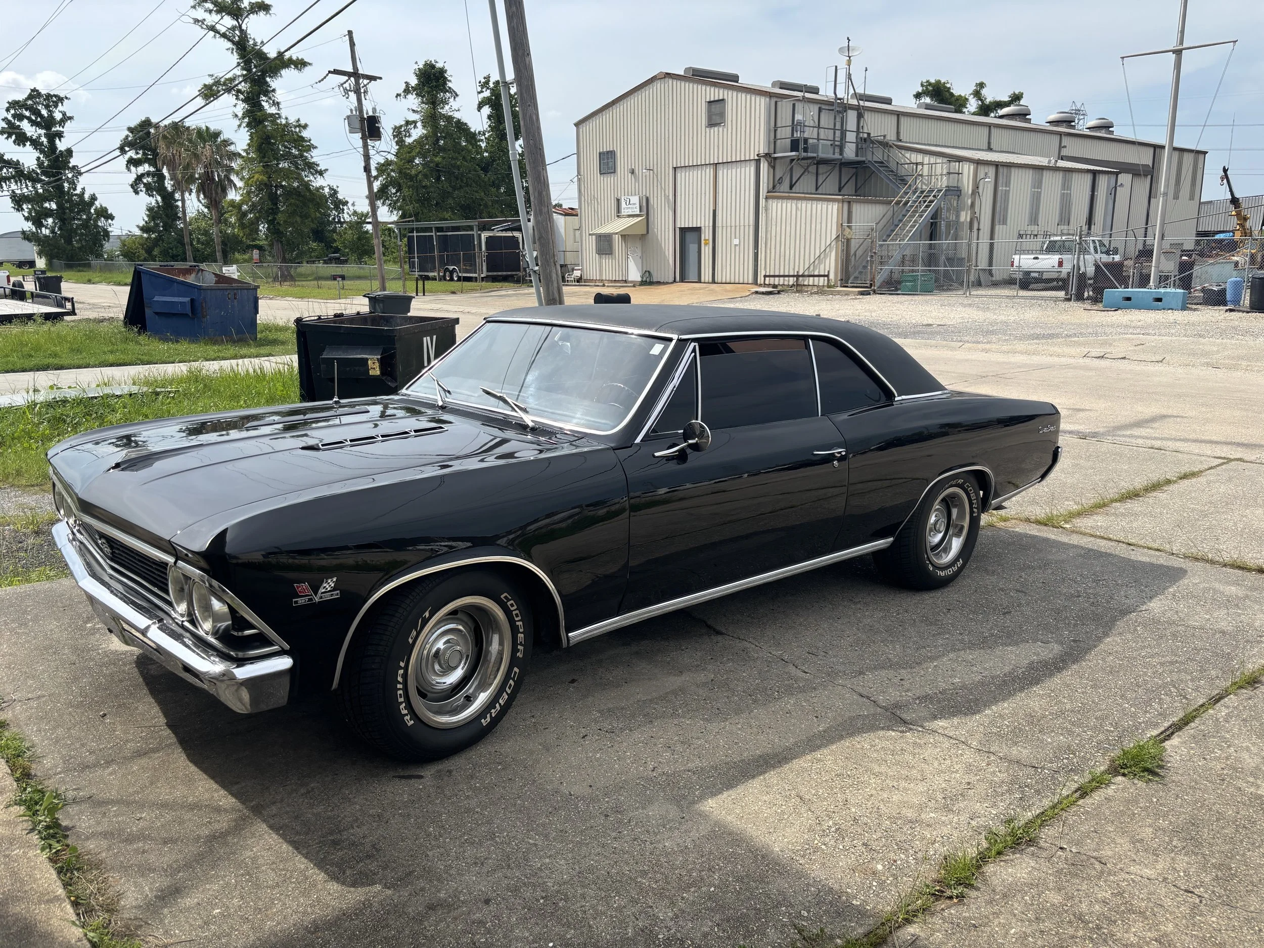 A black vintage car parked on a concrete lot with an industrial building and trees in the background.