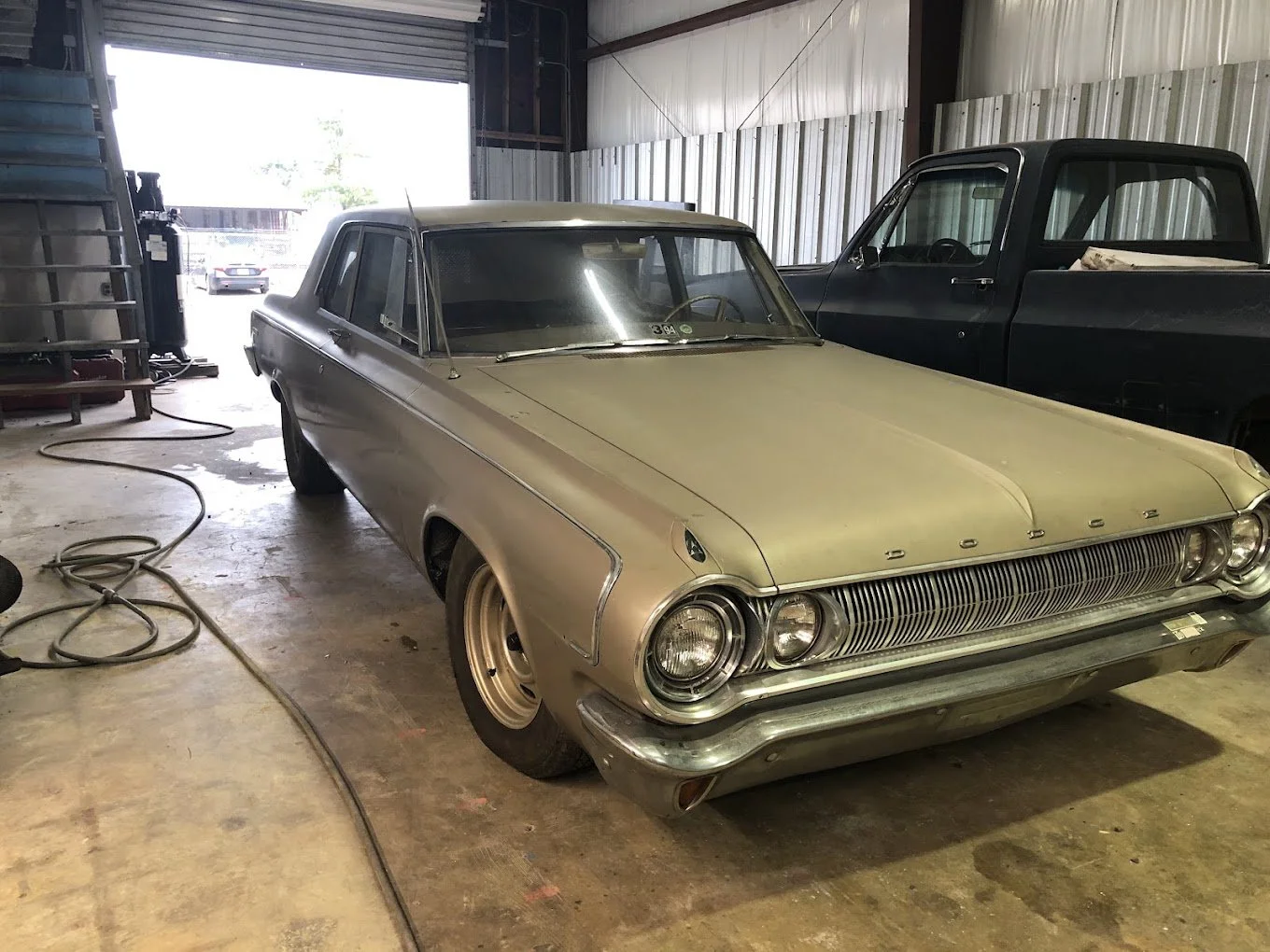 A vintage beige Ford car parked inside a garage, with another black truck and some equipment visible in the background.