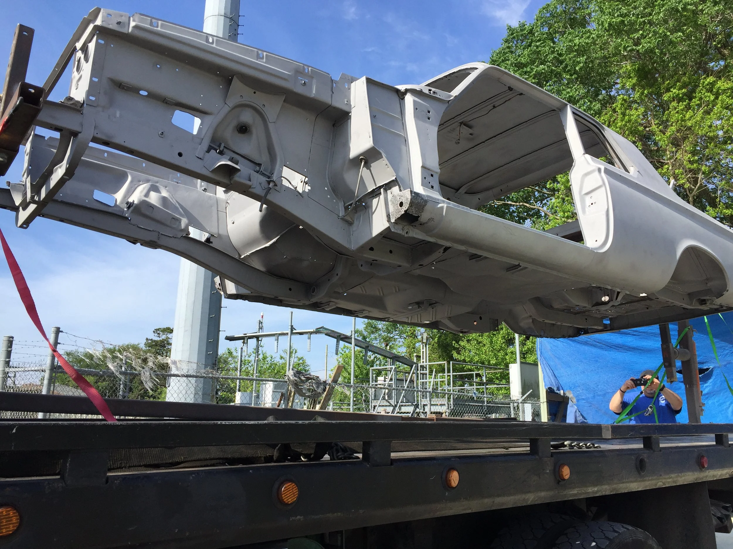 An unfinished white vehicle or truck body is mounted on a flatbed trailer, with a person taking a photo nearby. The sky is clear and blue, and there are green trees in the background.
