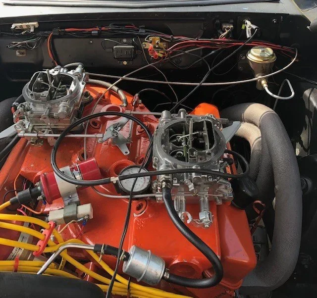 View of a car engine with orange engine block, carburetors, wiring, and various engine components.