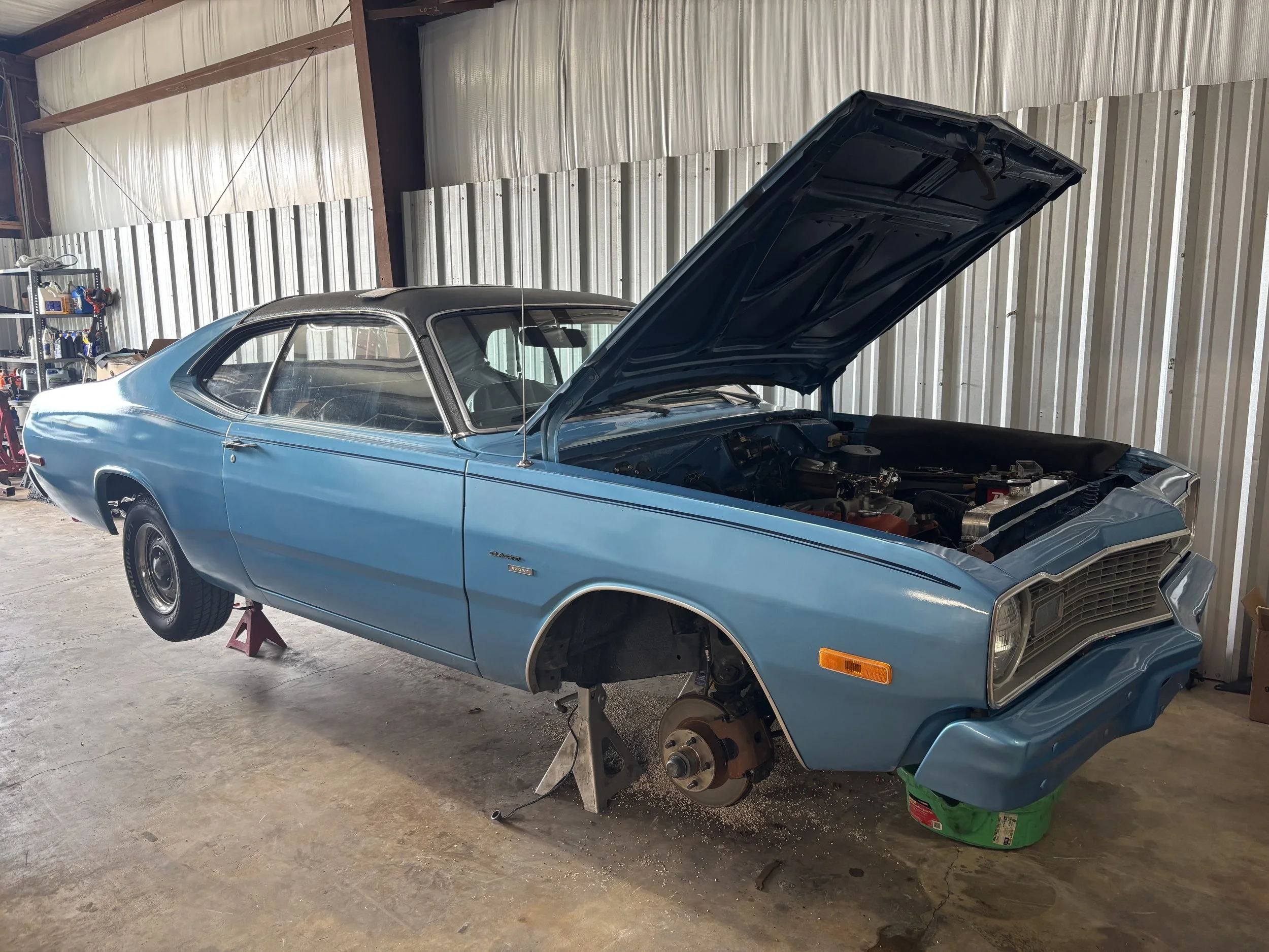 A vintage blue car is under maintenance in a garage with the hood open, elevated on jack stands, with no front wheels, and a workshop shelf in the background.