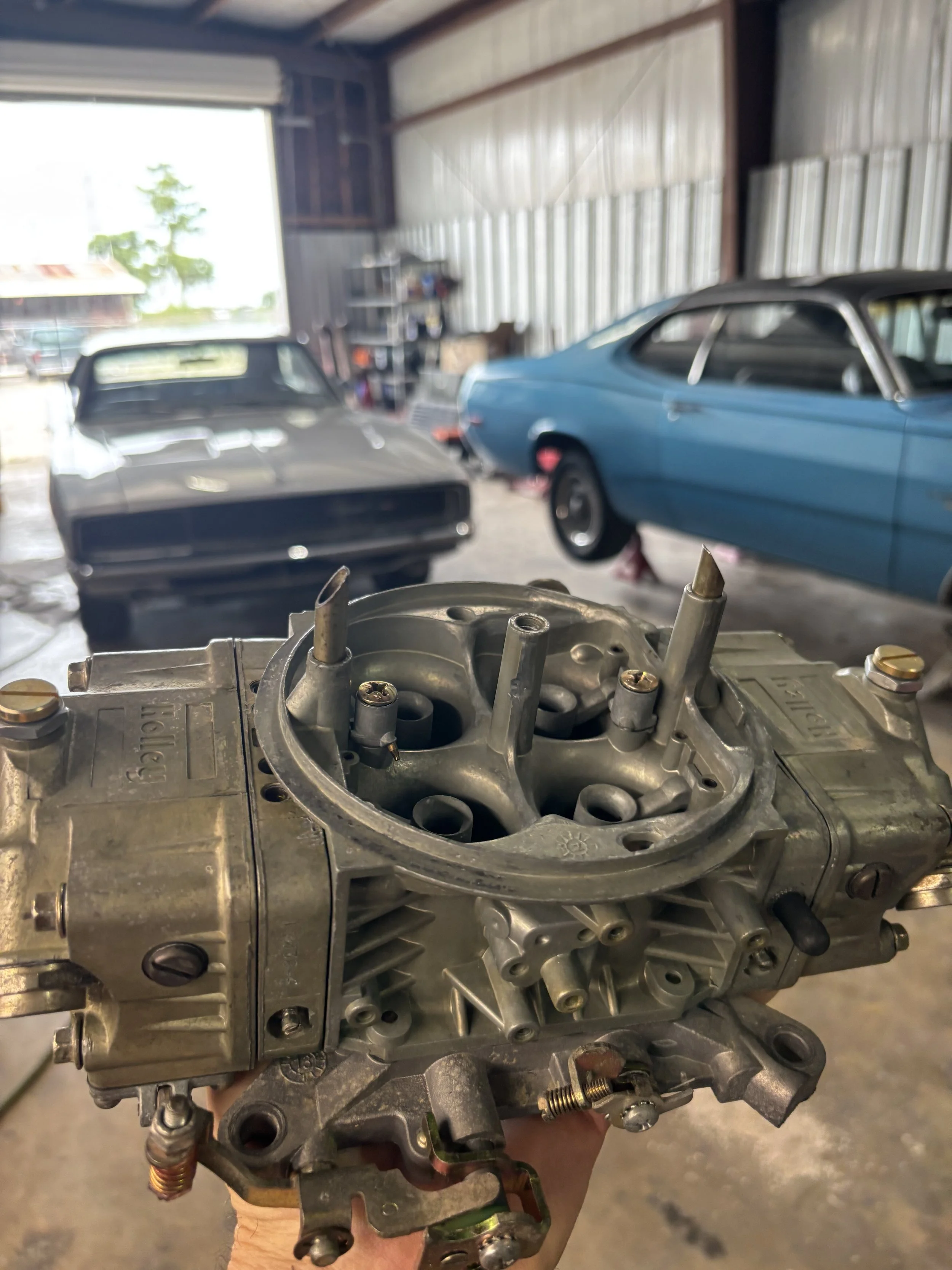 A car engine part is held up inside a garage with two classic cars, one black and one blue, visibly parked inside.