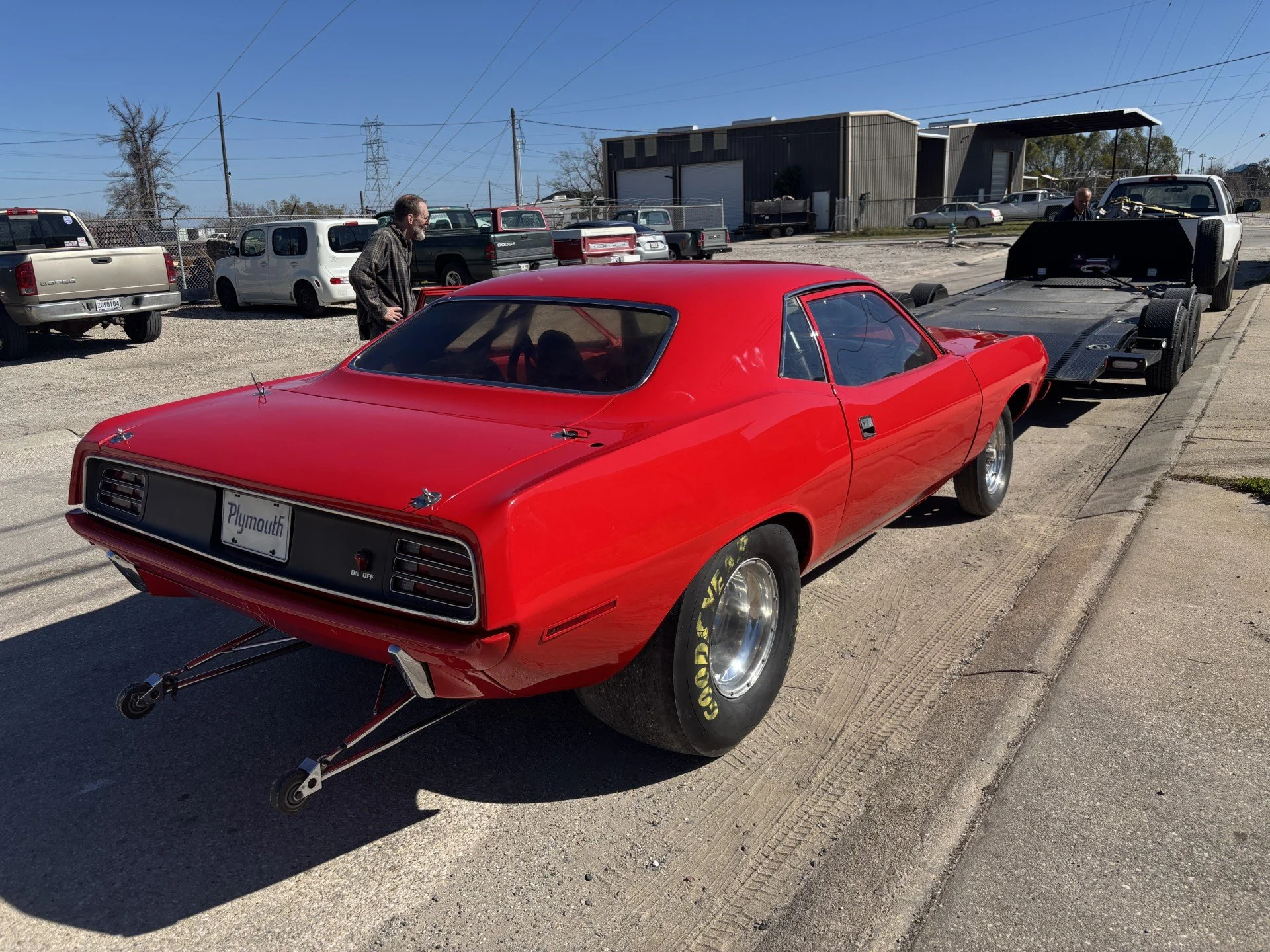 A red vintage Plymouth muscle car with racing tires parked on the street, with a street scene and several trucks and cars in the background, and two men inspecting the cars.