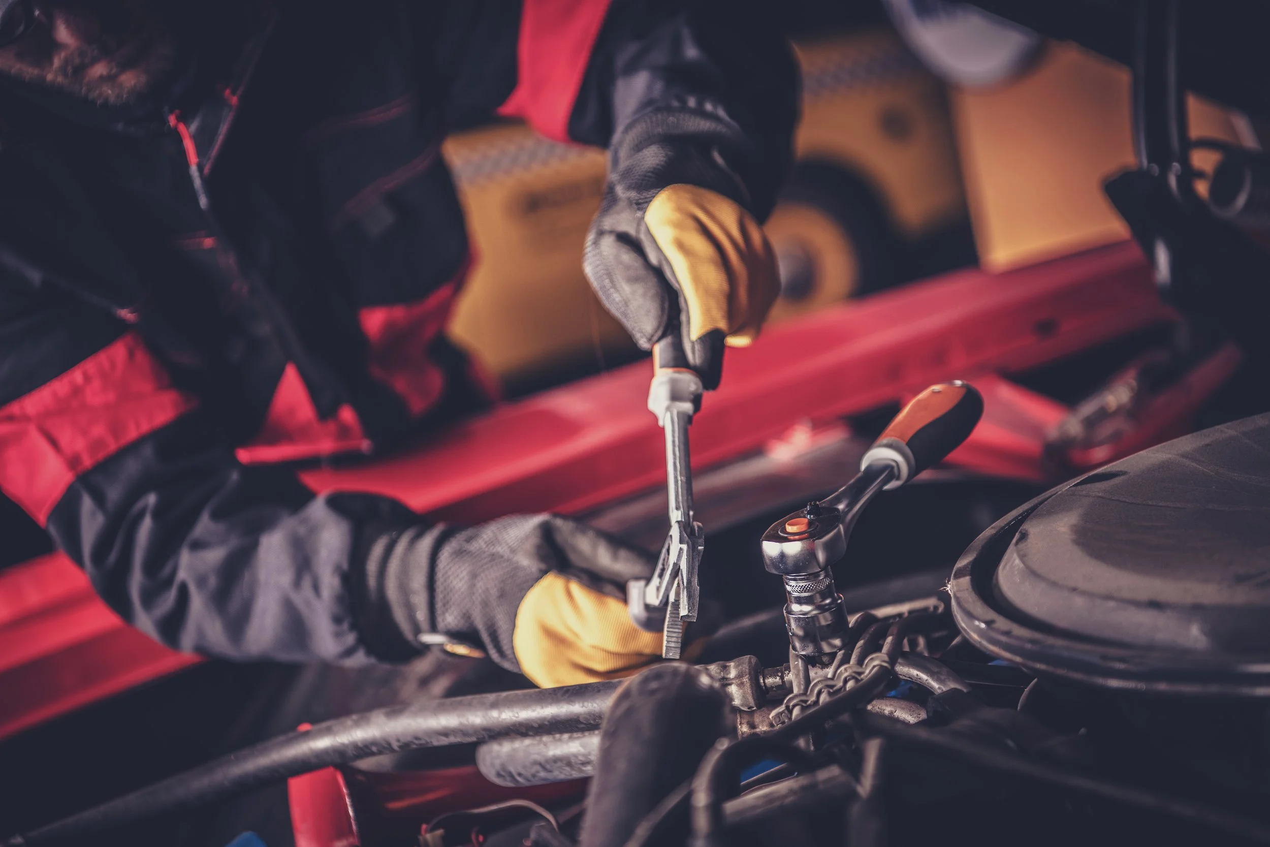 A mechanic wearing yellow gloves working on a motorcycle engine inside a workshop with tools and equipment in the background.