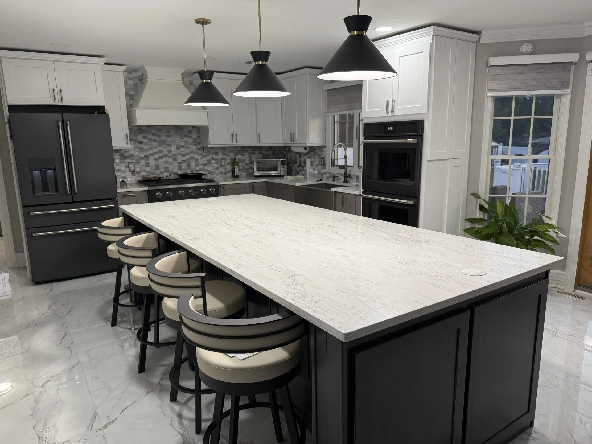 Modern kitchen with white cabinets, black appliances, and a marble island with bar stools. Features gray tile backsplash, large windows, and pendant lighting.