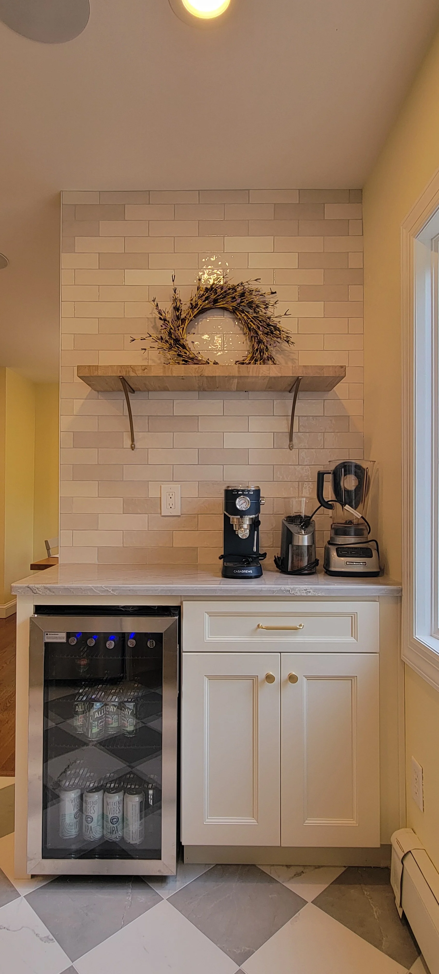 A small kitchen counter with a coffee maker, an electric kettle, a blender, and a small wine fridge. Above the counter is a wooden shelf decorated with a wreath, mounted on a beige brick wall. A yellow wall is partially visible on the right side.