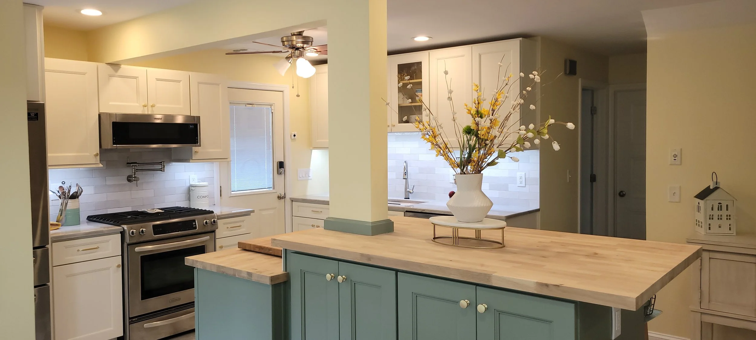 A modern kitchen with white cabinets, a stainless steel stove, microwave, and refrigerator. A blue kitchen island with a wooden countertop holds a white vase with yellow and white flowers. There is a ceiling fan with lights, a window, and a door with blinds.