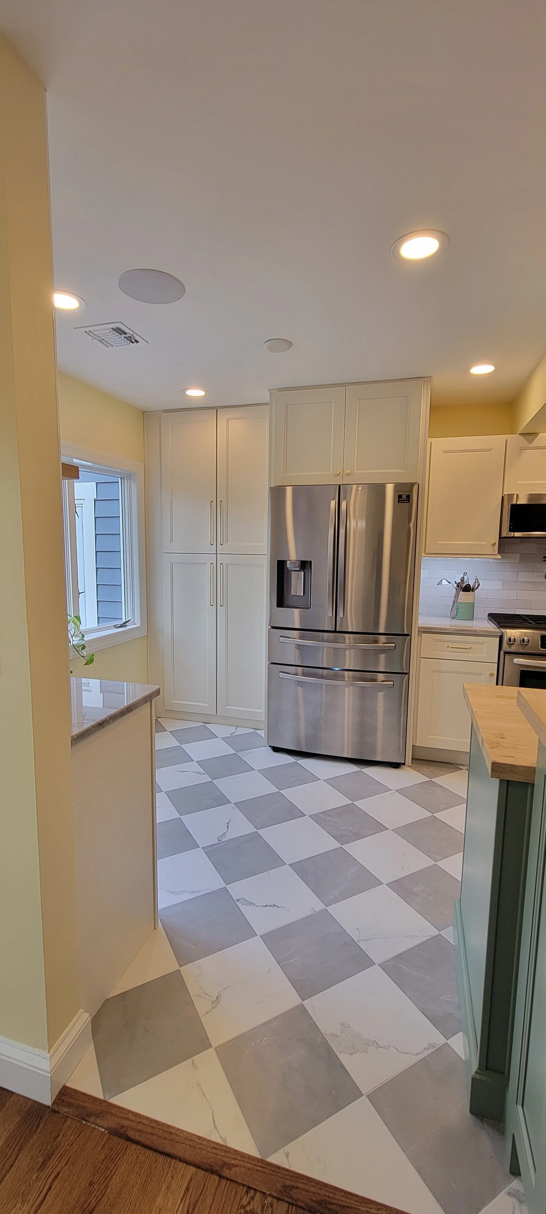 A modern kitchen with white cabinets, a stainless steel refrigerator, a window, and checkered gray and white floor tiles.