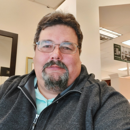 A man with glasses, a beard, and curly hair taking a selfie in an indoor setting, possibly an office.