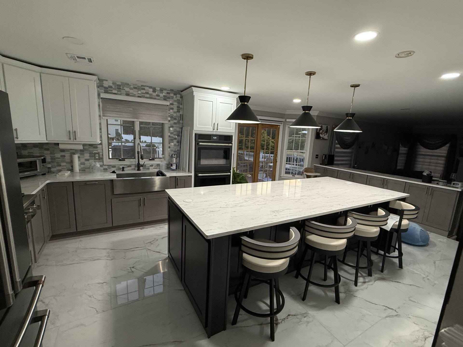 Modern kitchen with white and gray cabinetry, marble floor, and a large white island with black accents and barstools. Pendant lighting above.
