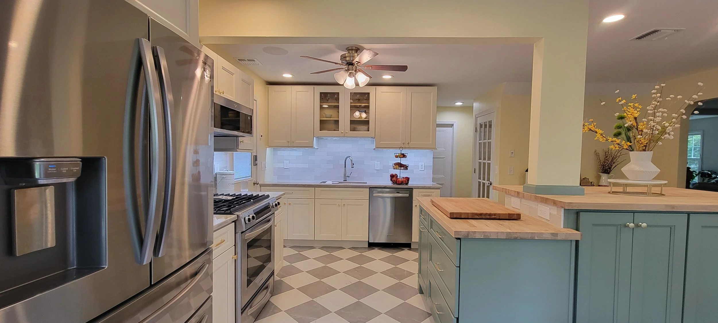 Modern kitchen with white cabinetry, stainless steel appliances, a light green island with a wooden countertop, a white tiled backsplash, and decorative items like a flower arrangement and a fruit basket.