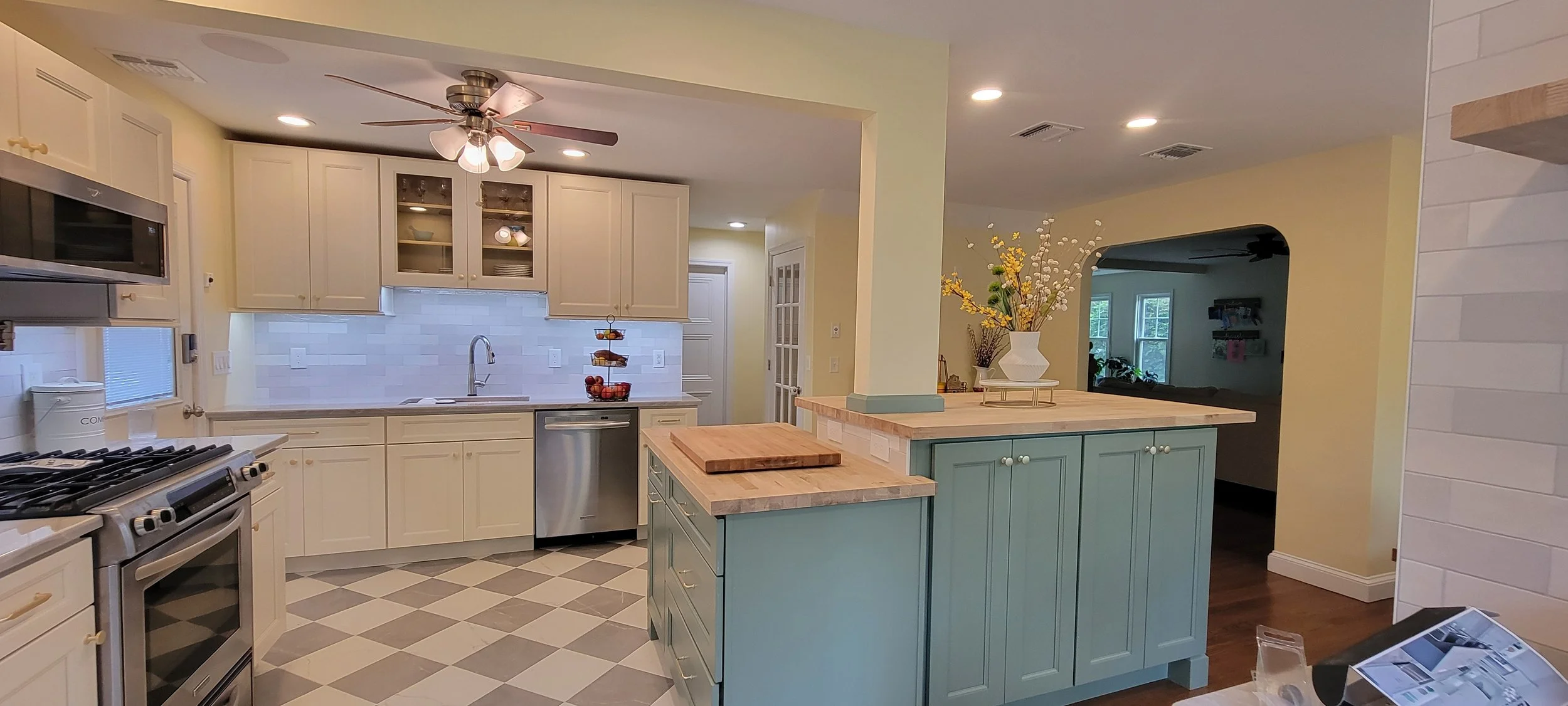 A bright kitchen with white cabinets, a blue island with a wooden top, and a white tile backsplash. There is a ceiling fan with lights, a stainless steel dishwasher, and a fruit basket on the counter. A decorative vase with yellow and white flowers is on the island.