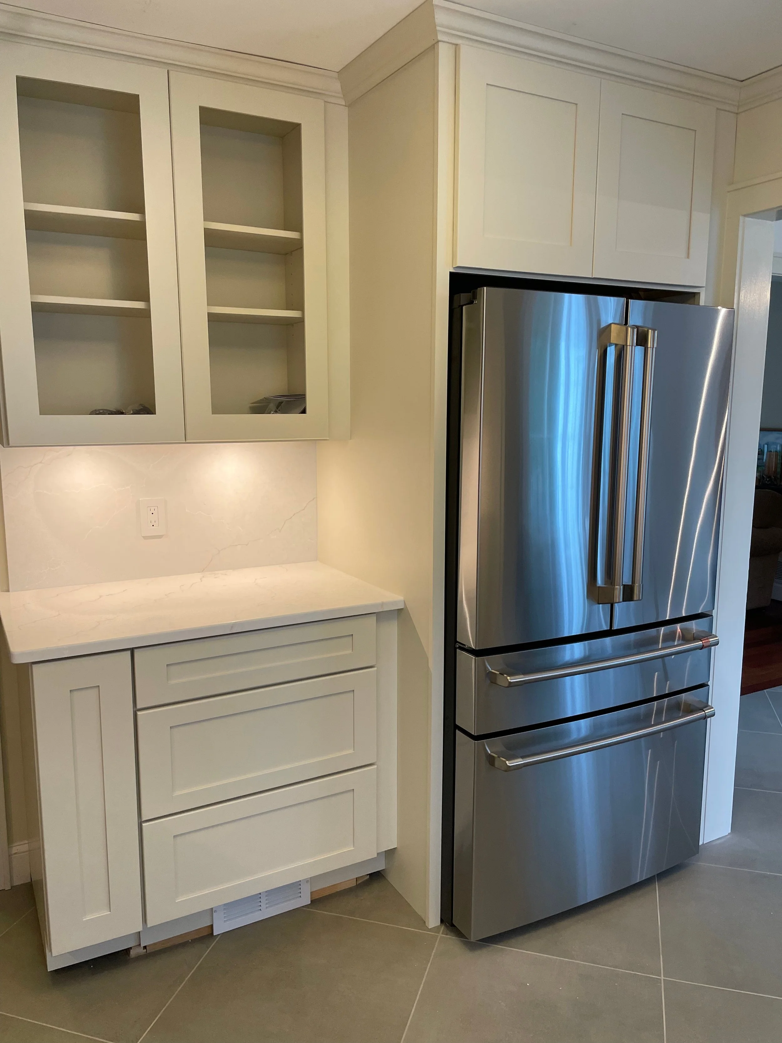 Kitchen corner with white cabinets, open shelf cabinet, and a stainless steel refrigerator with bottom drawers on a tiled floor.