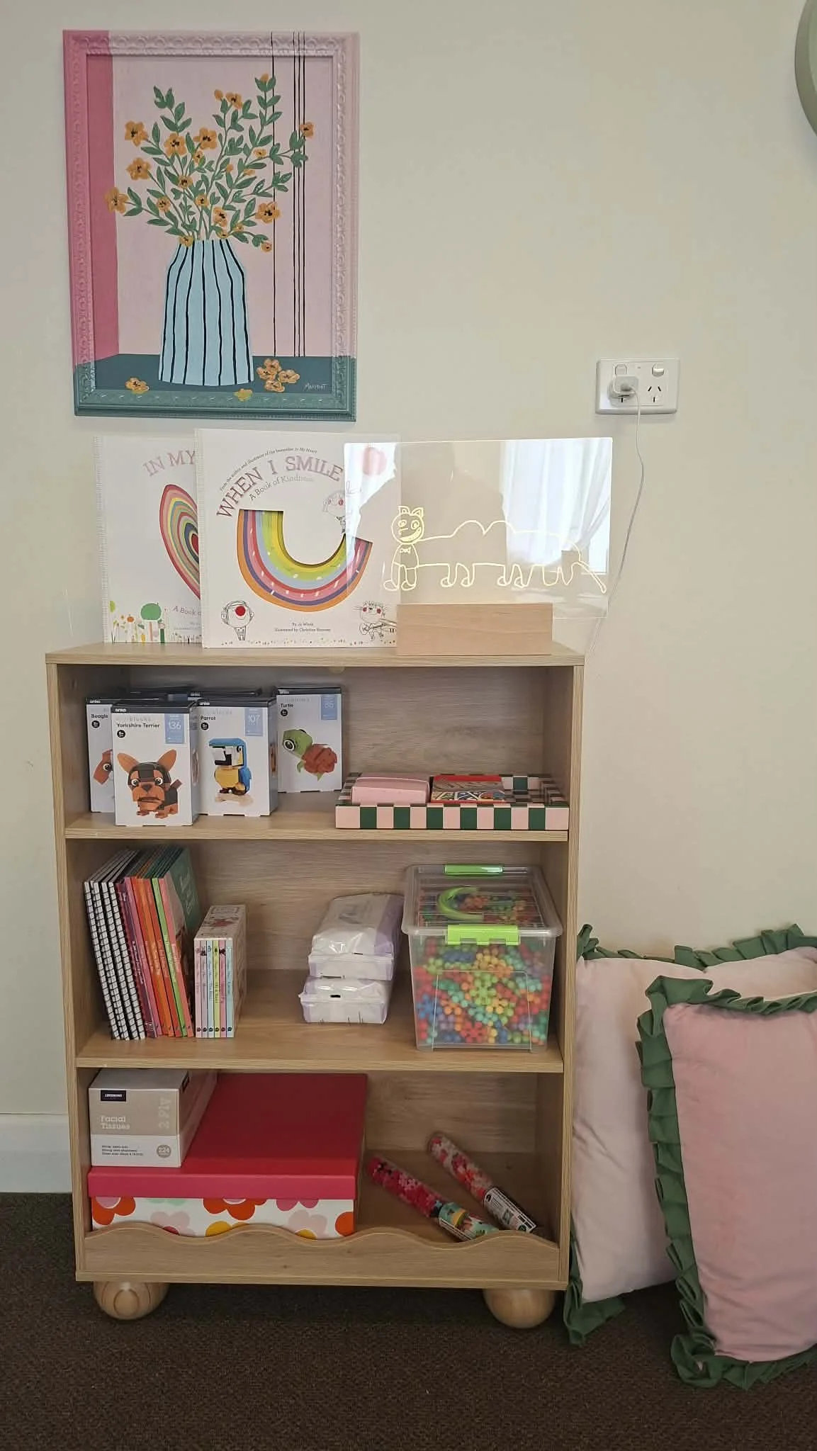 A wooden bookshelf filled with children's toys, books, and games, next to a pink and green beanbag chair, with a wall decorated with a framed painting of a blue striped vase with yellow flowers, and a neon light outline of a lion and other animals.