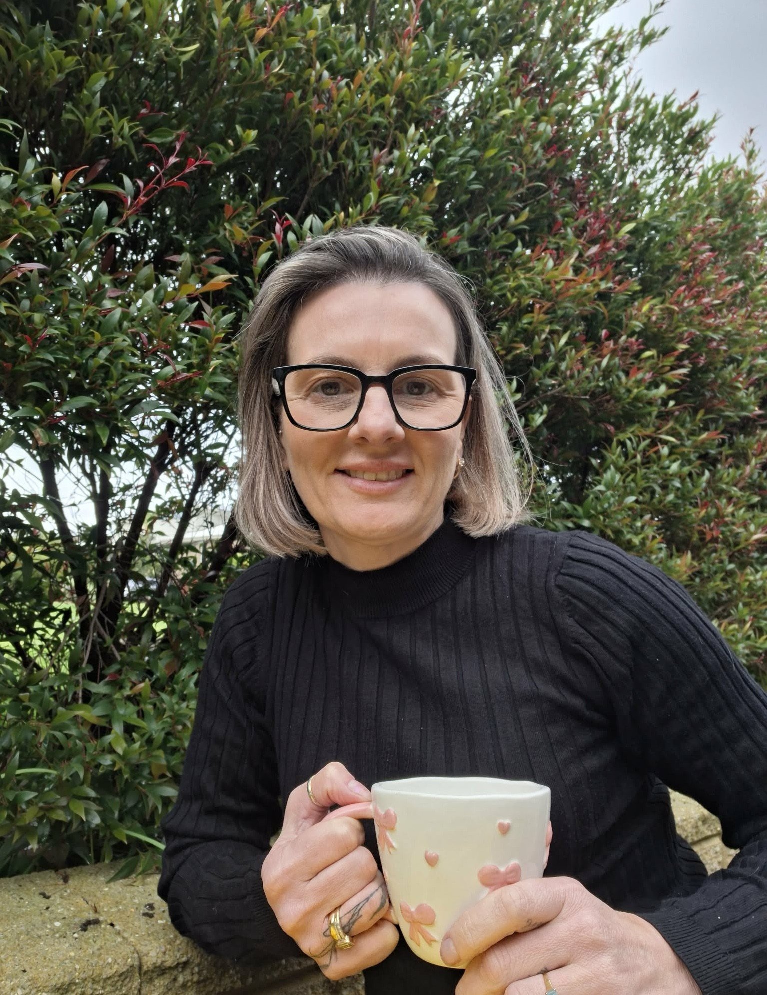 A woman with shoulder-length gray hair, wearing glasses and a black long-sleeve top, smiling while holding a white mug decorated with pink hearts. She is sitting outdoors in front of a bush with green and reddish leaves, and holding the mug with both hands.