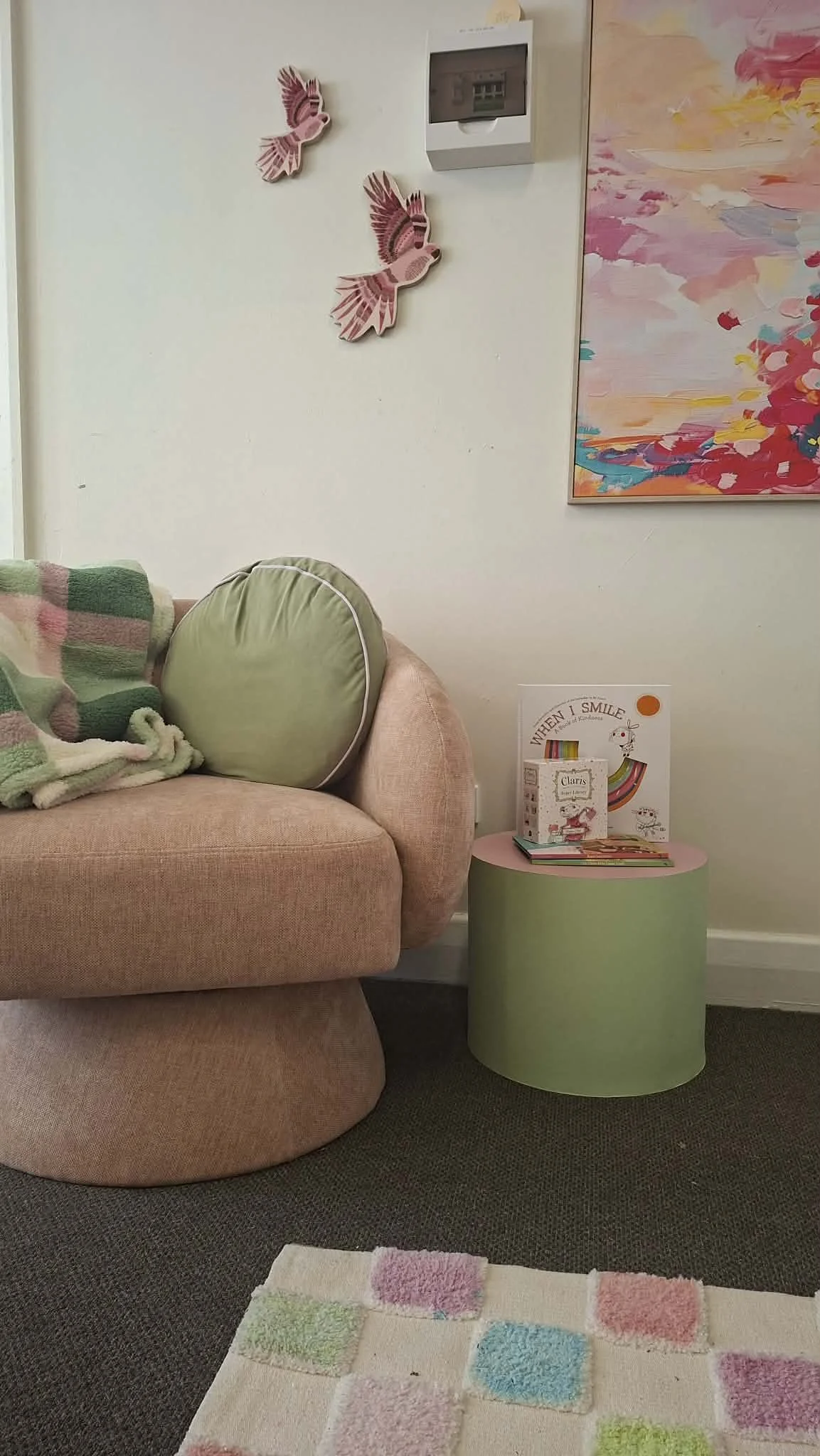 A pink upholstered armchair with a round green pillow and a pastel-colored blanket. A round green side table holding children's books and a framed picture. Wall decor includes two pink bird decorations and a colorful abstract painting.