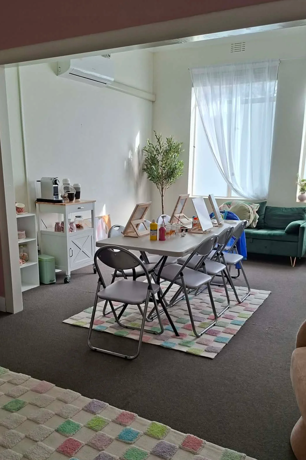 A cozy living room with a dining table set up with art supplies, a sofa, a large window with sheer curtains, a potted plant, and a colorful rug.