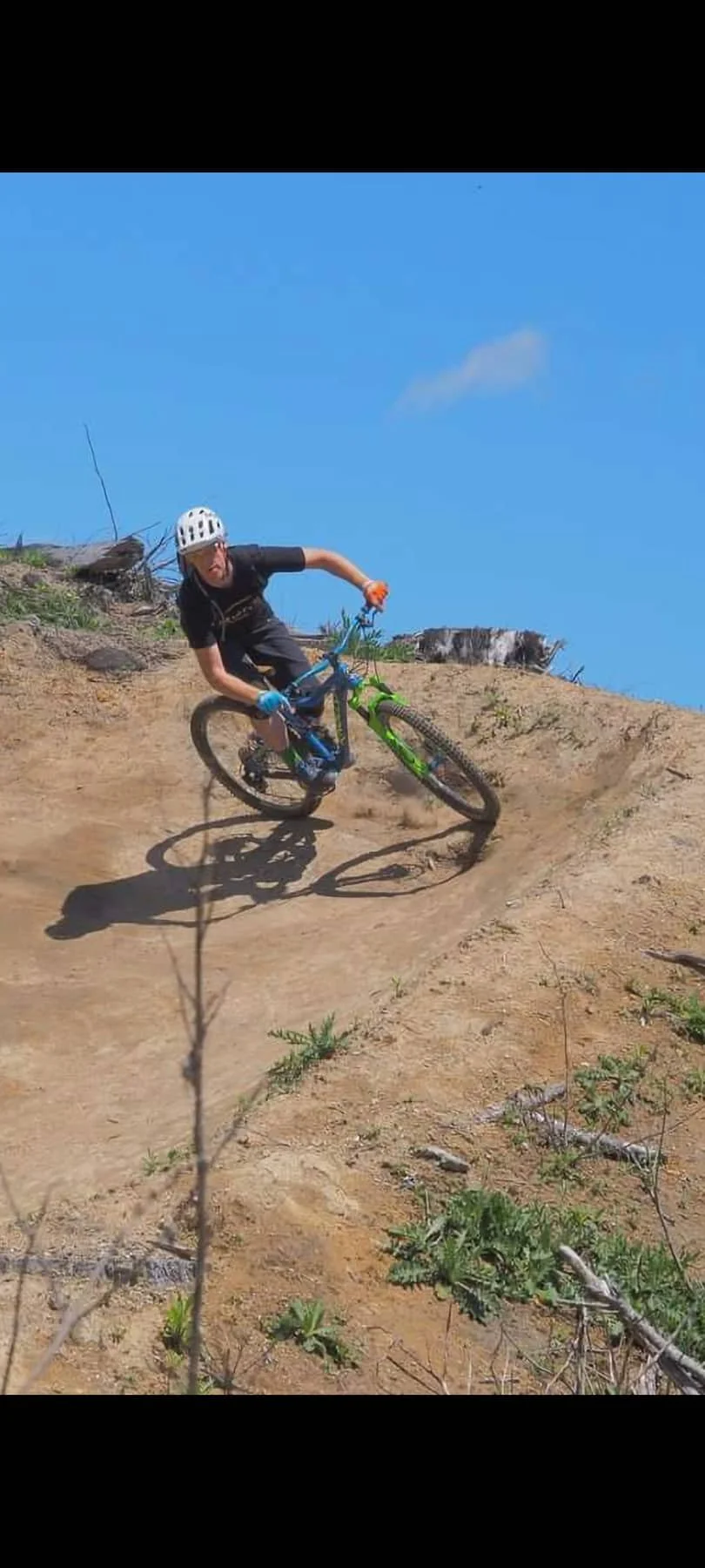 A person riding a mountain bike downhill on a dirt trail, wearing a helmet, black shirt, blue gloves, and black shorts, under a clear blue sky.