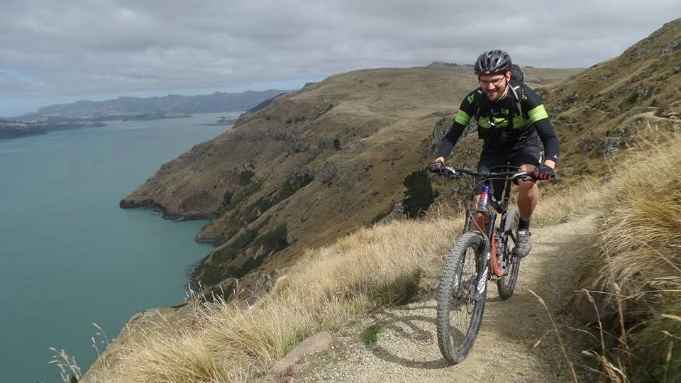 A person riding a mountain bike on a narrow dirt trail along a hillside with a view of a body of water and rolling hills in the background.