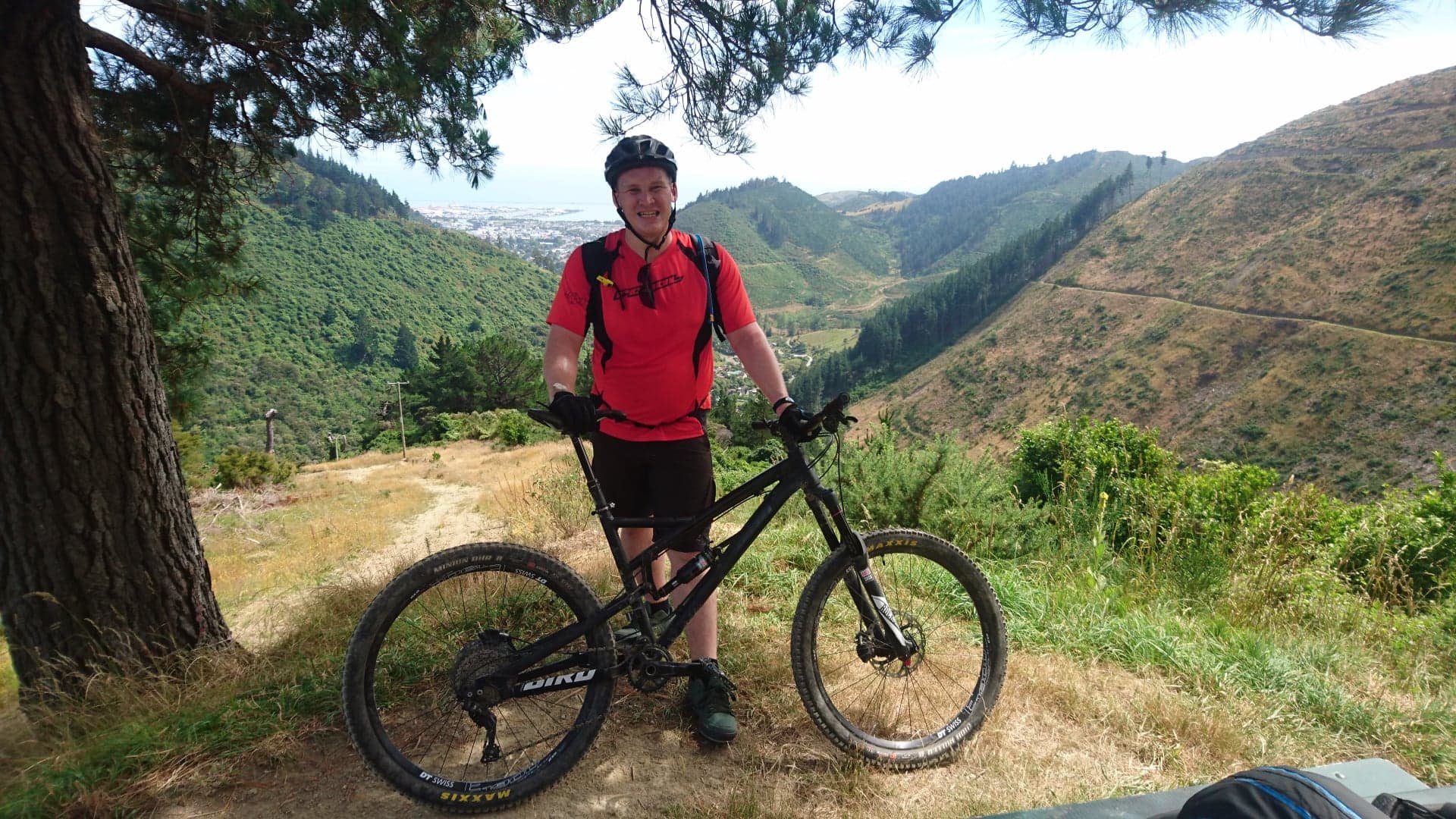 A person in a red shirt and helmet standing with a mountain bike on a trail in a hilly, green landscape, with mountains and a town visible in the background.