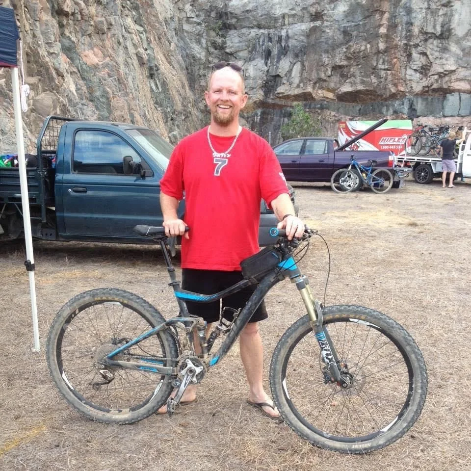A man in a red shirt and black shorts standing outdoors with a mountain bike, smiling at the camera. The background shows trucks, bikes, and a rocky cliff face.