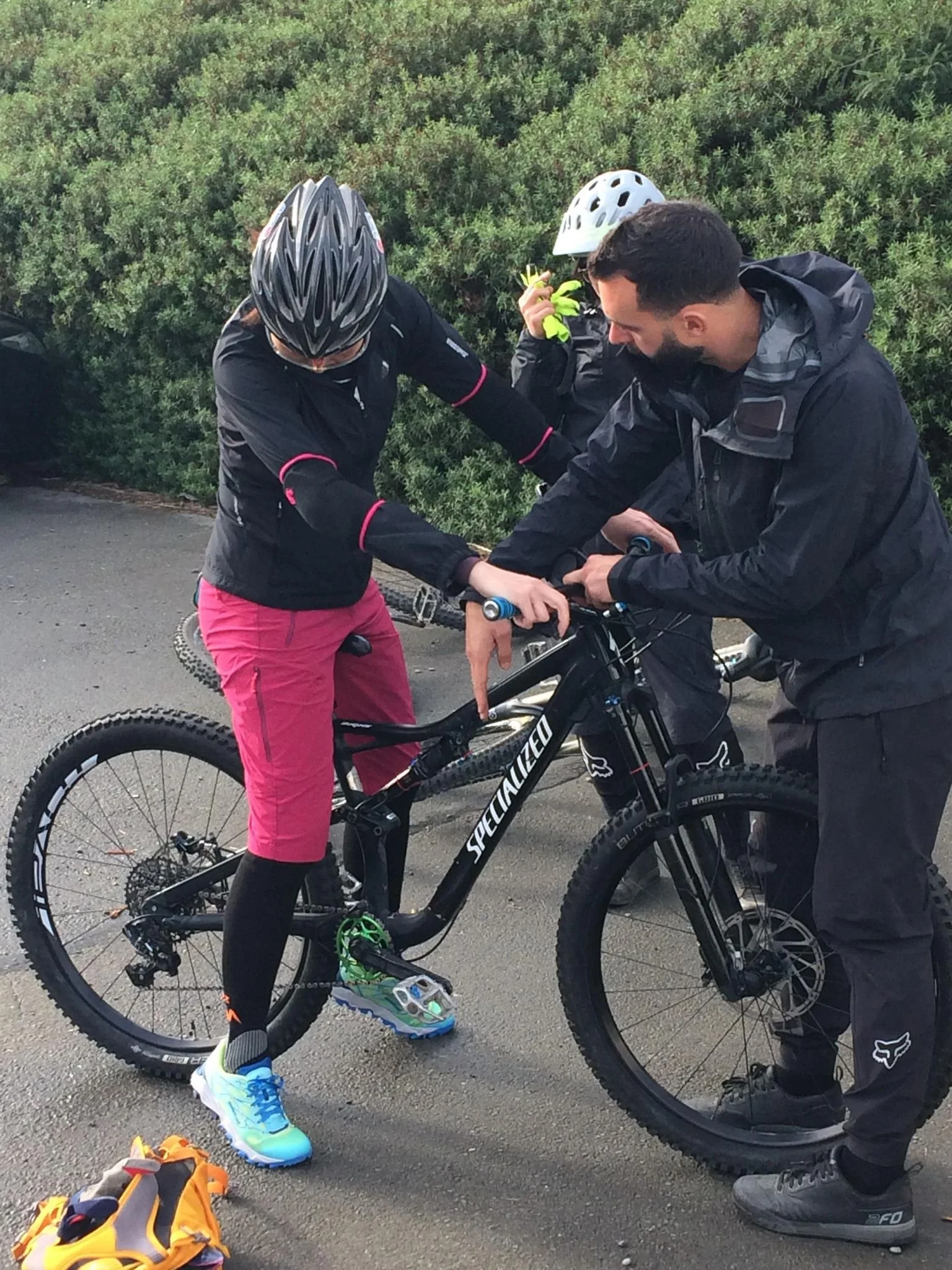 Two people with helmets and athletic clothing preparing their bicycles, with a bushy green background and a yellow bag with cycling gear on the ground.