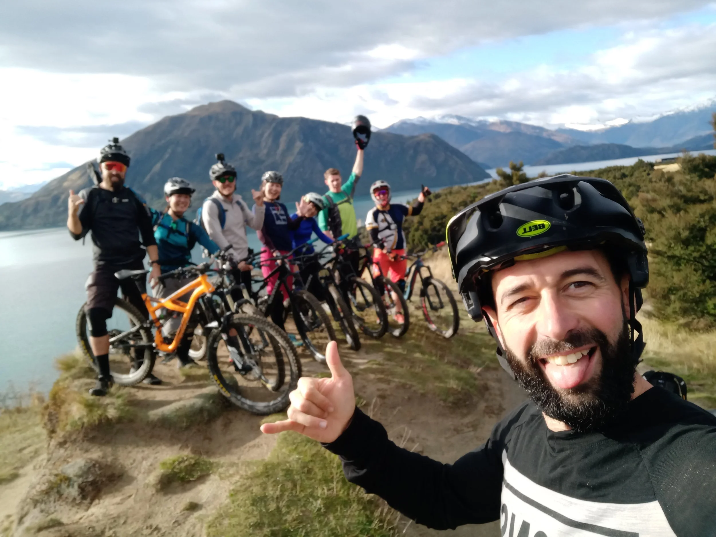 A group of six mountain bikers standing with their bikes near a lake, with mountain peaks in the background, and a man in the foreground taking a selfie with friends.
