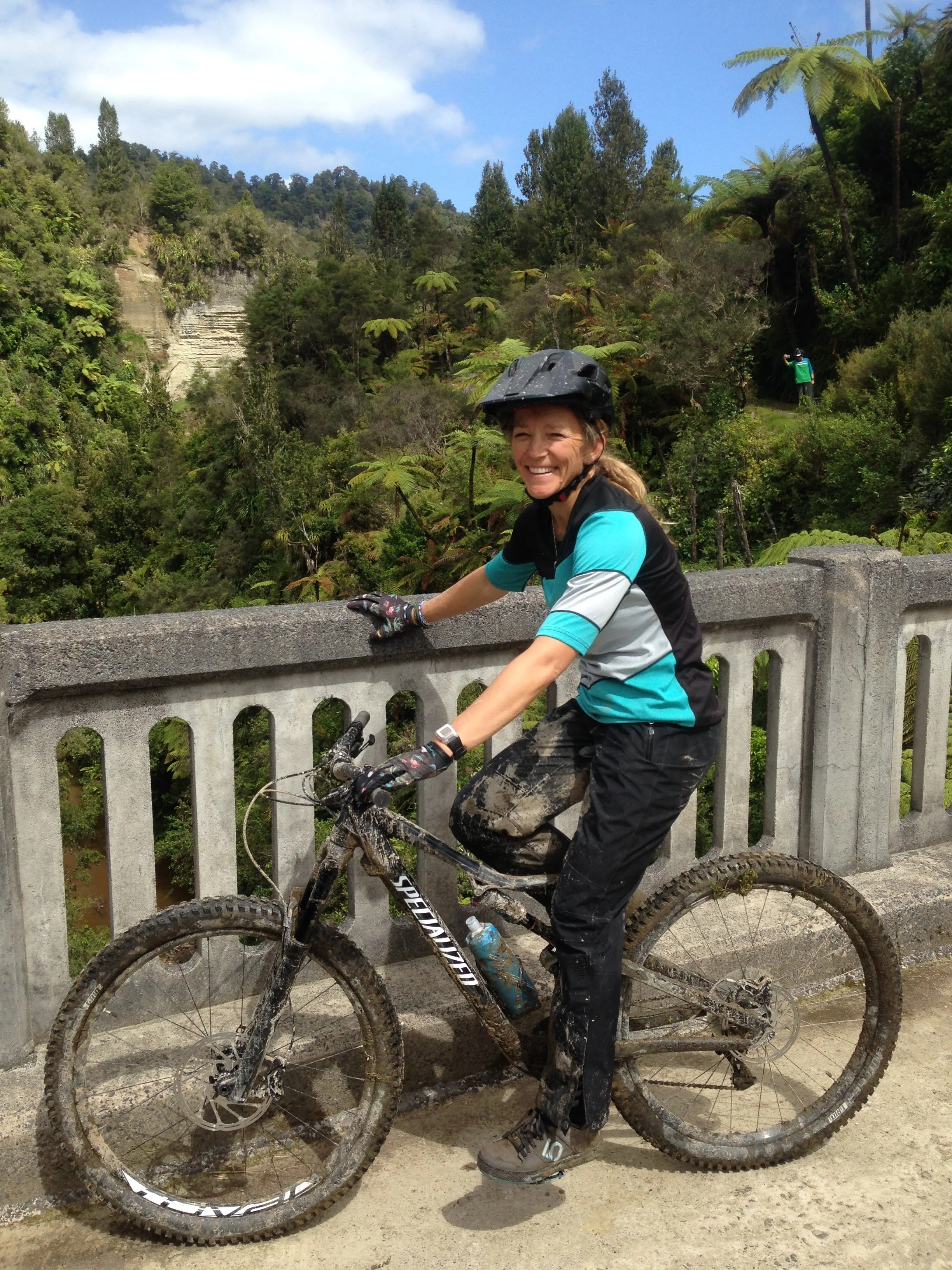 A woman in a helmet and cycling gear smiling next to her muddy mountain bike, standing on a bridge with a lush forest and mountains behind her.