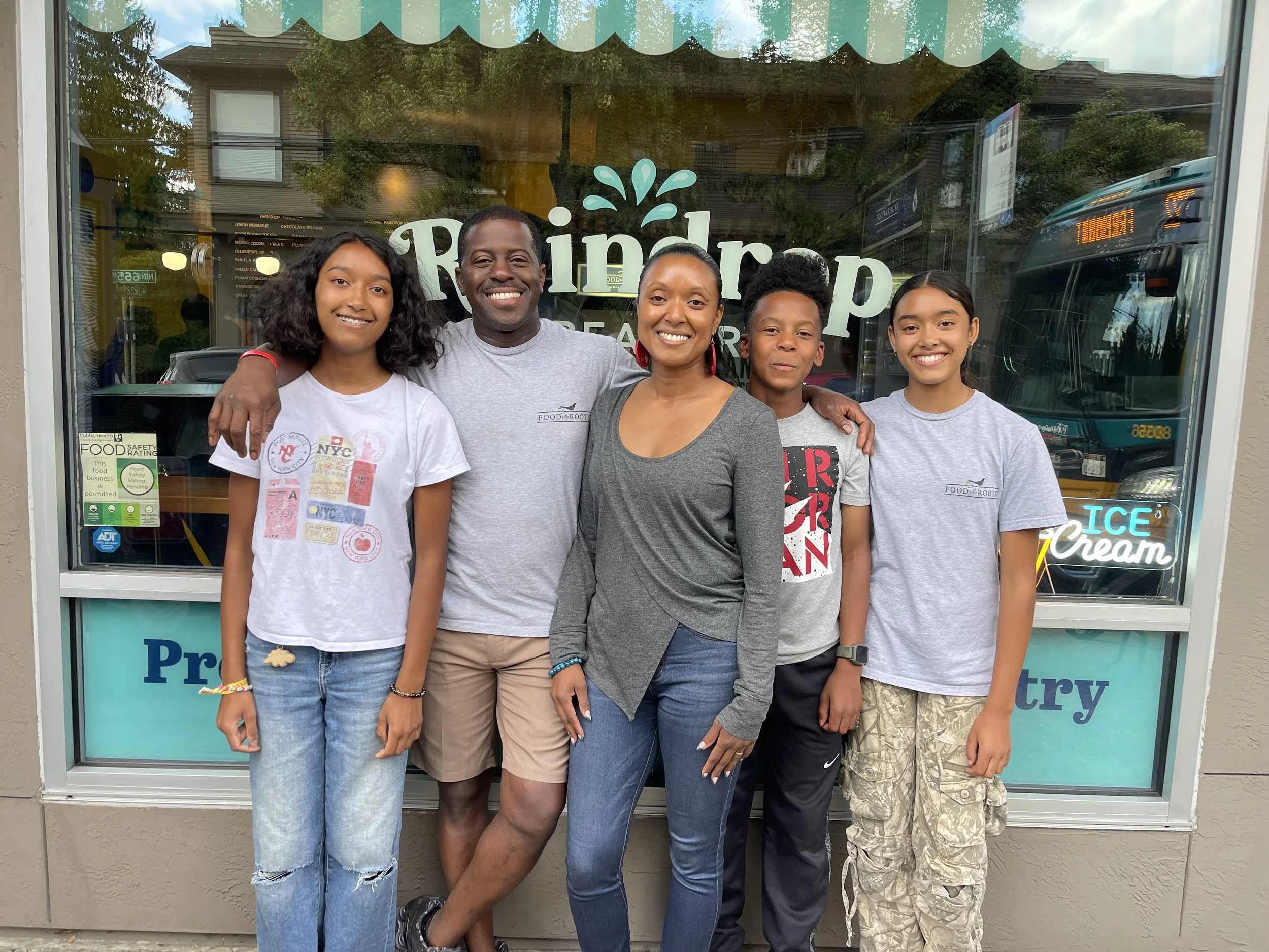Owner and family standing outside in front of a storefront window. The group includes two adults and three children, all smiling and posing for the photo.