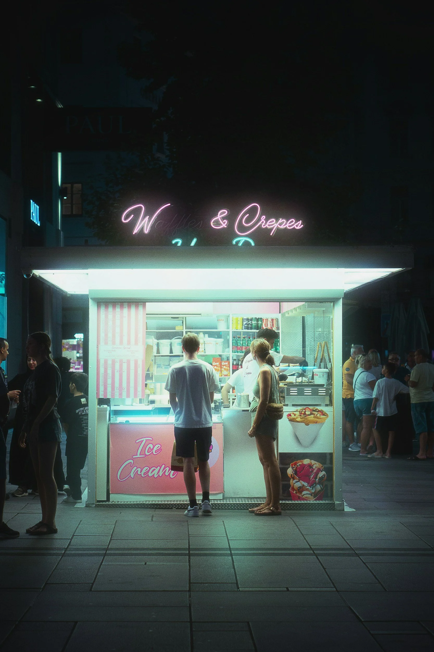 Nighttime scene of an ice cream stand with neon pink and green signs that read 'Waffles & Crepes' and 'Ice Cream'. People are gathered around, some ordering or waiting for ice cream or waffles, with a city sidewalk and pedestrians in the background.