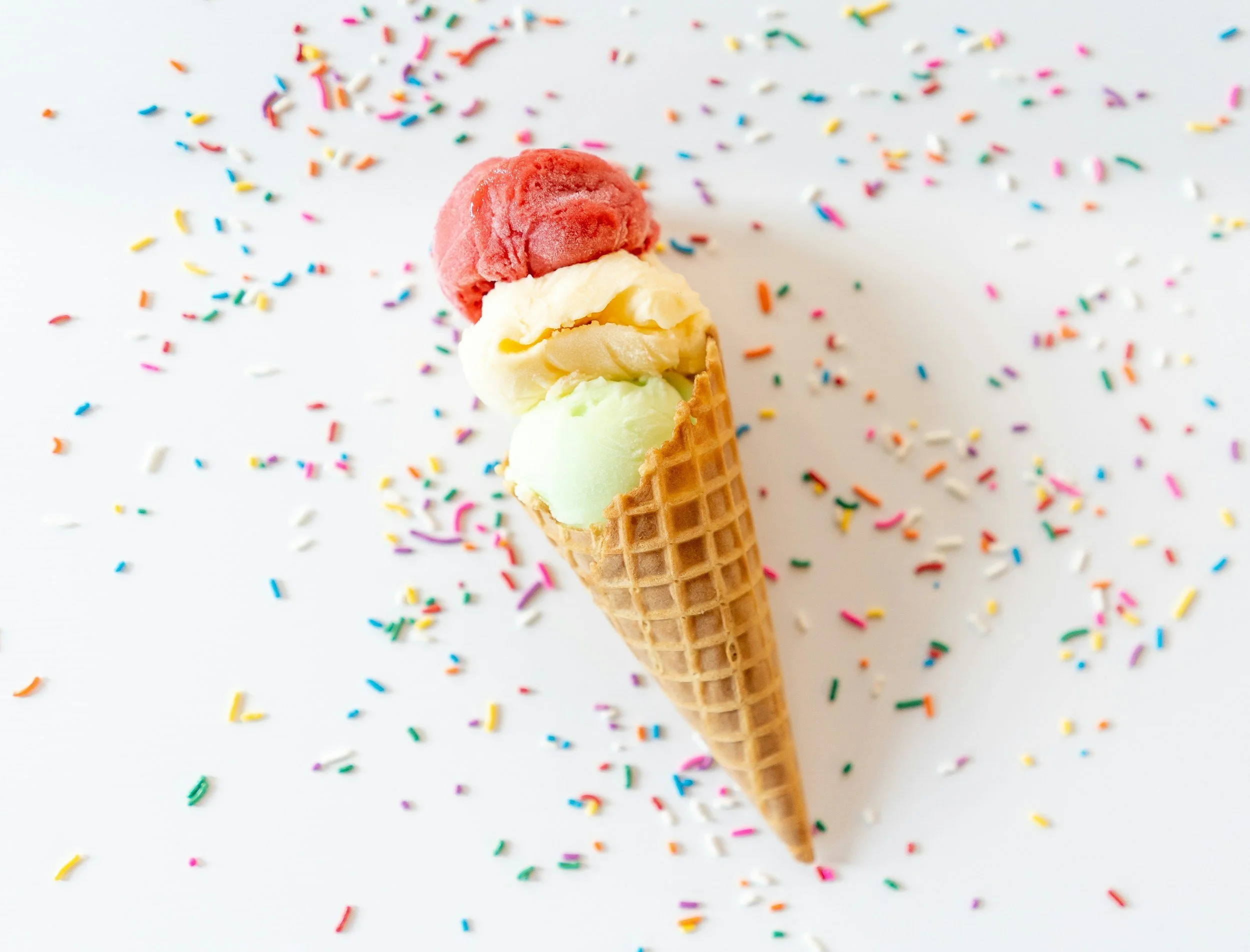 Three scoops of ice cream in a waffle cone with rainbow sprinkles scattered on a white surface.