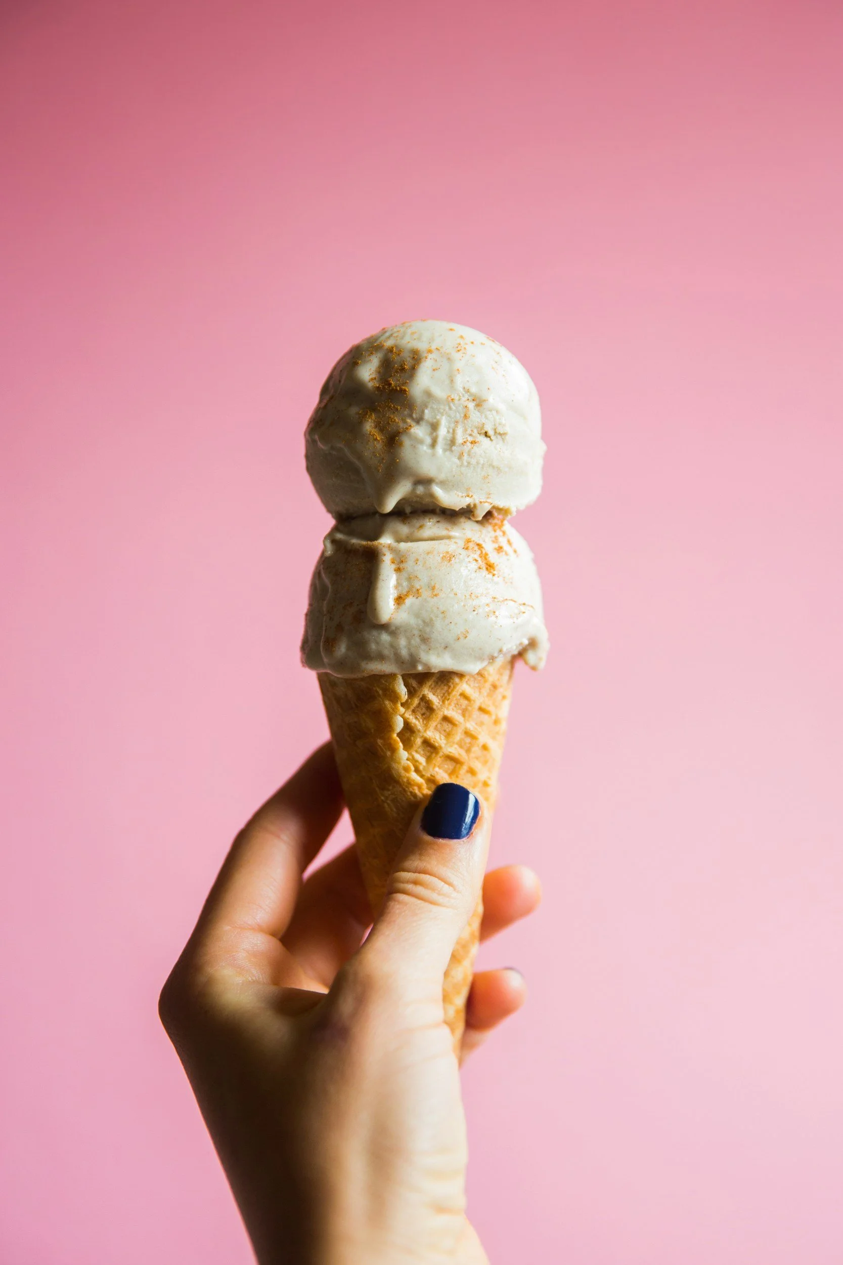 A hand holding a double scoop ice cream cone with vanilla ice cream topped with cinnamon, against a pink background.
