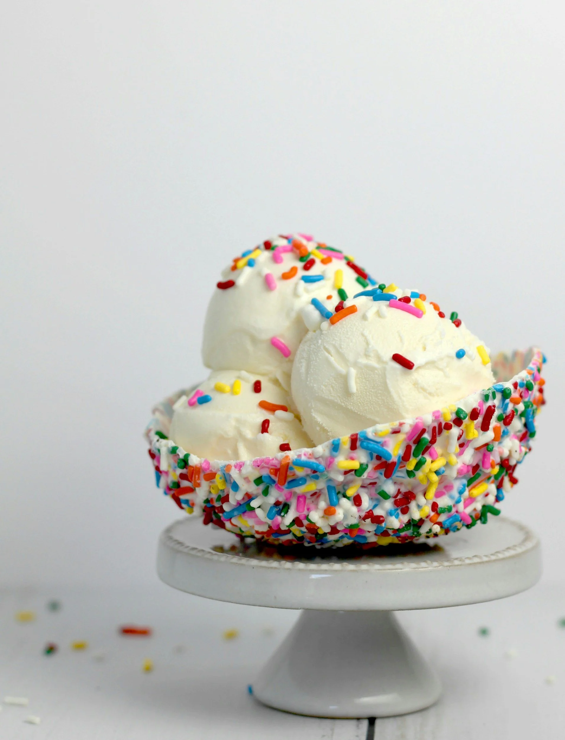 Three scoops of vanilla ice cream topped with colorful rainbow sprinkles in a small dish on a white surface with loose sprinkles scattered around.