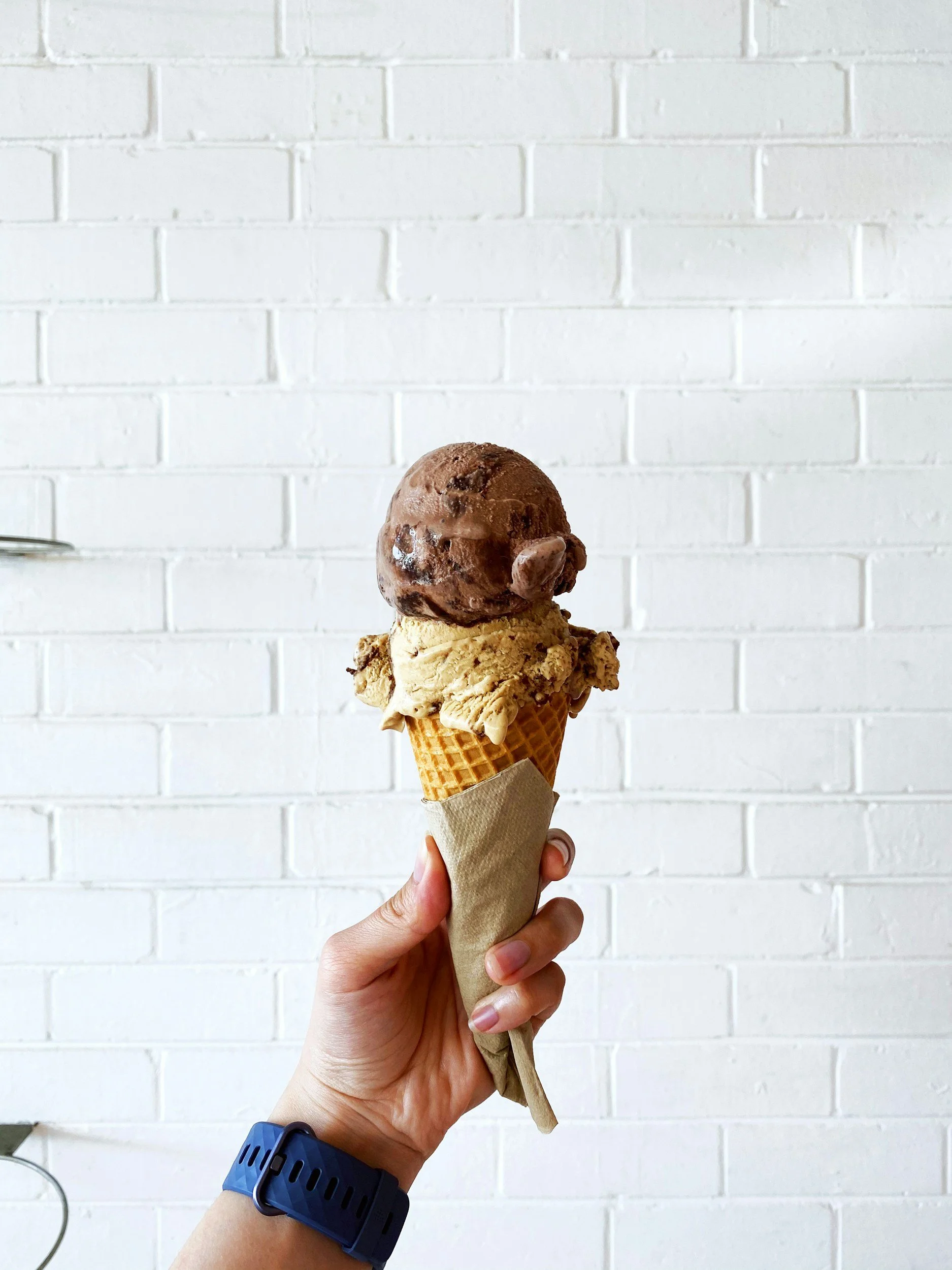 Person holding an ice cream cone with a scoop of chocolate ice cream against a white brick wall background.