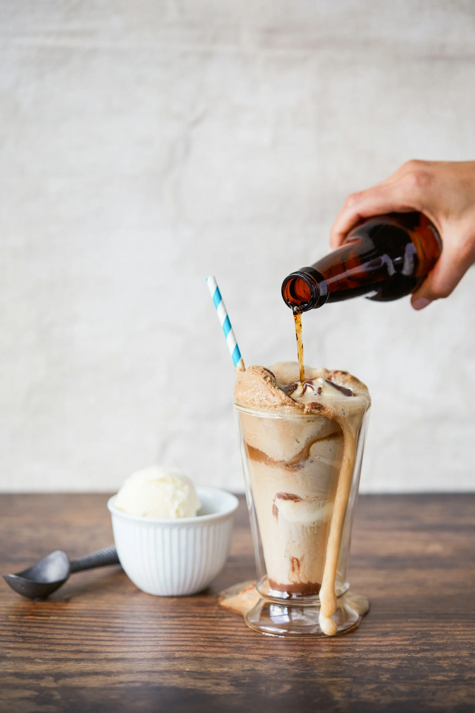 A tall glass of ice blended caramel coffee topped with whipped cream and caramel syrup, with a hand pouring cola into it, on a wooden table. A bowl of vanilla ice cream and a spoon are in the background.