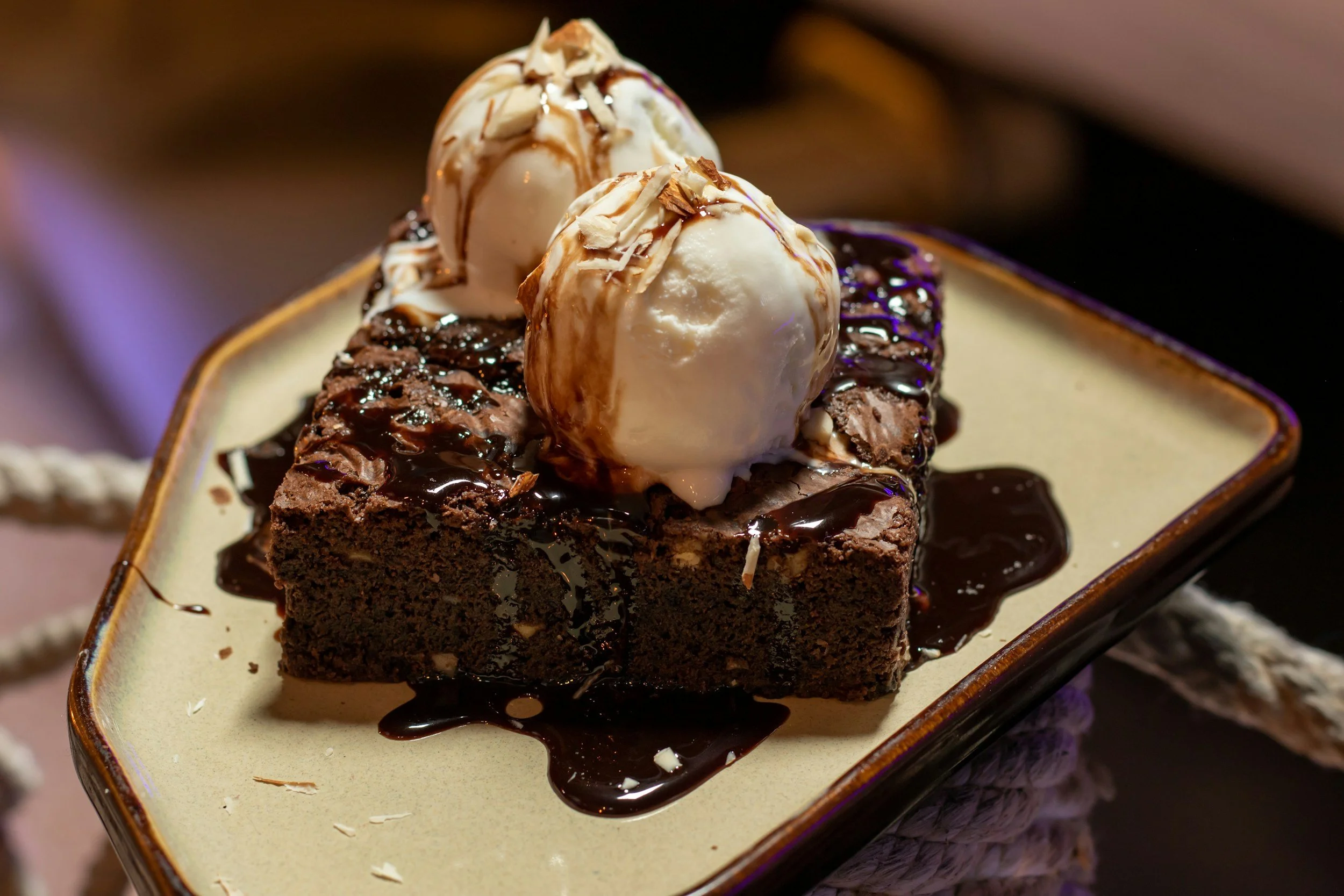 A chocolate brownie topped with vanilla ice cream scoops, drizzled with chocolate and caramel syrup, garnished with shaved white chocolate, served on a decorative ceramic plate.