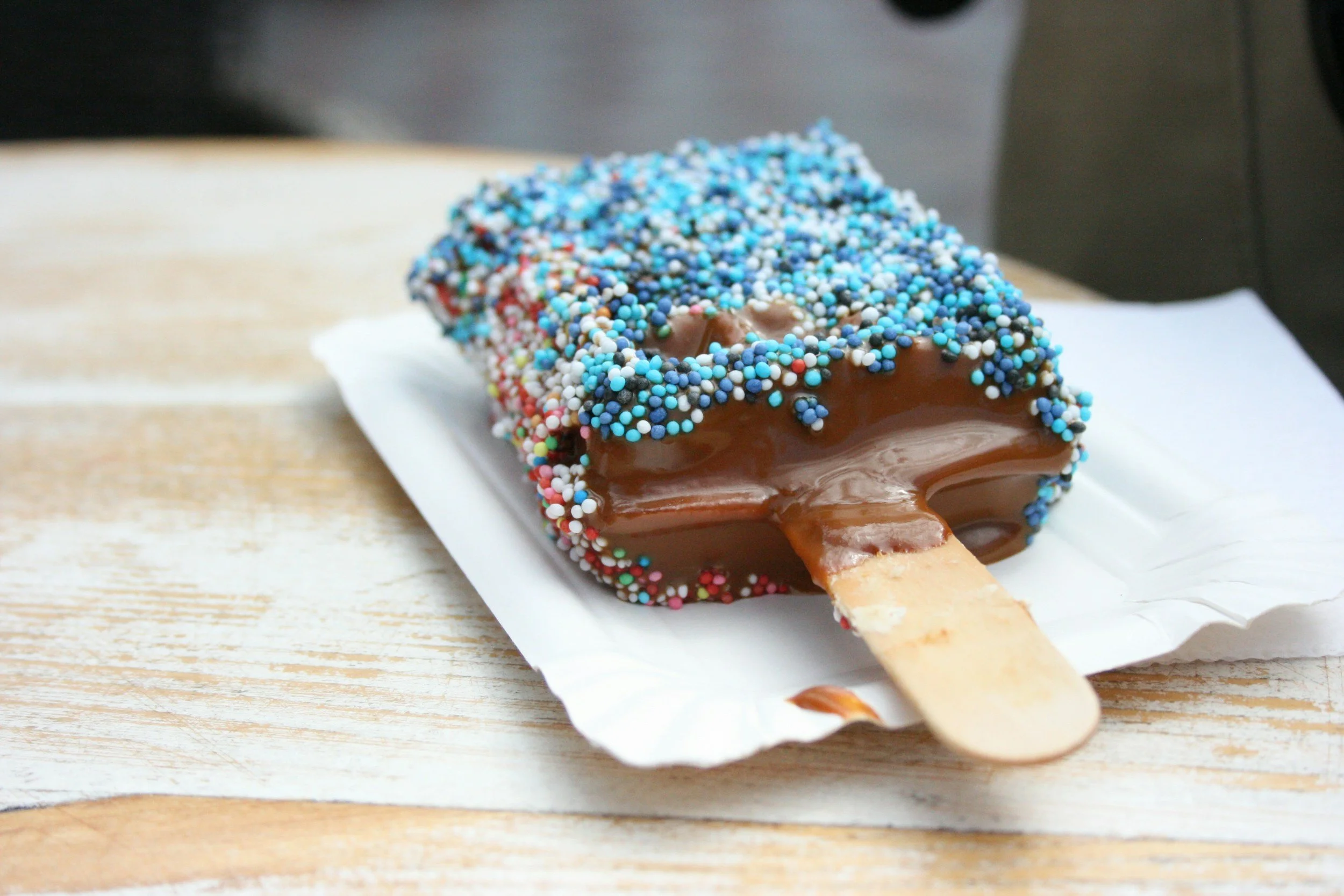 Chocolate ice cream bar coated with multicolored sprinkles on a wooden surface.