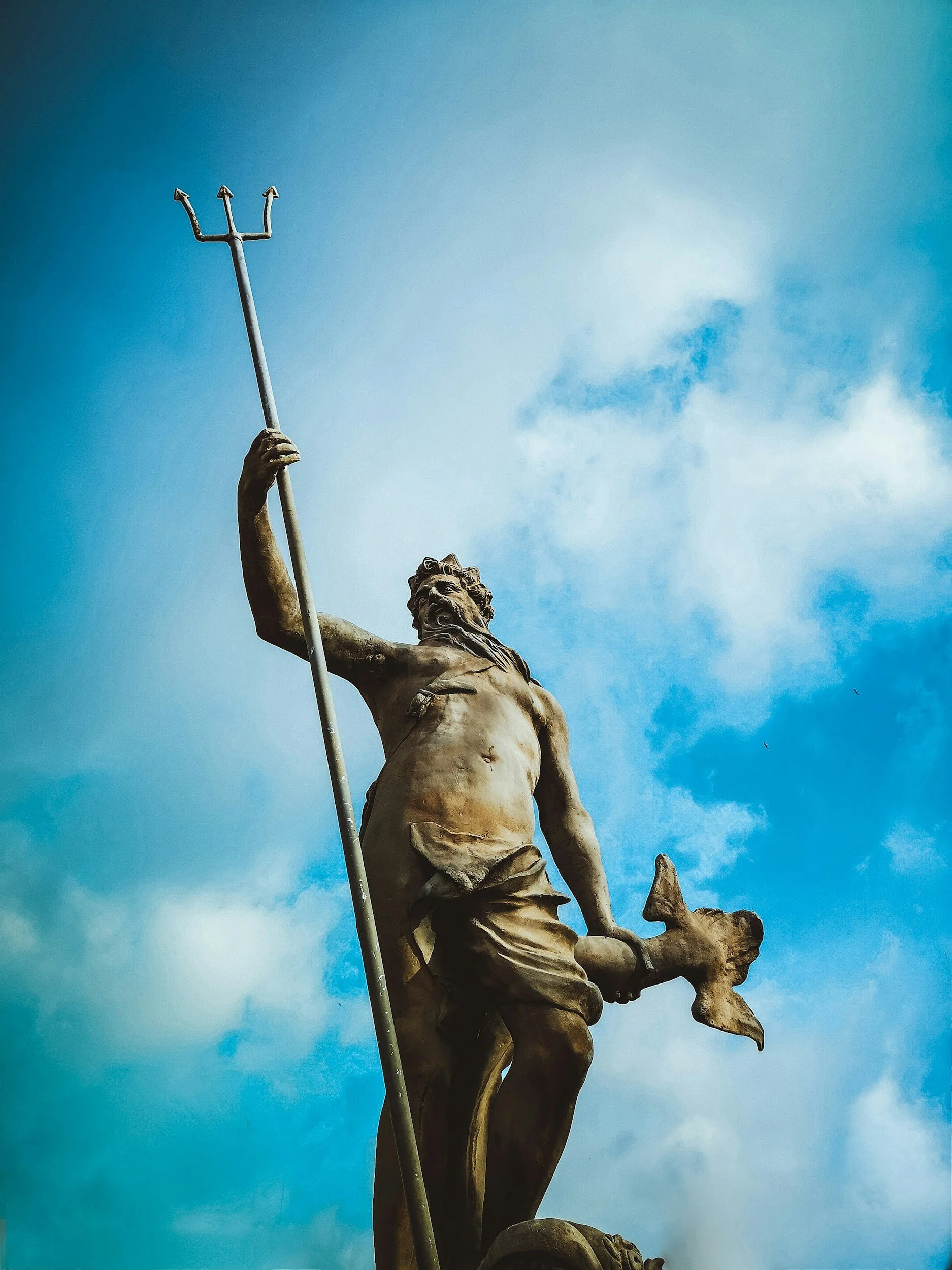 Bronze statue of a mythological figure holding a trident, with a blue sky and some clouds in the background.
