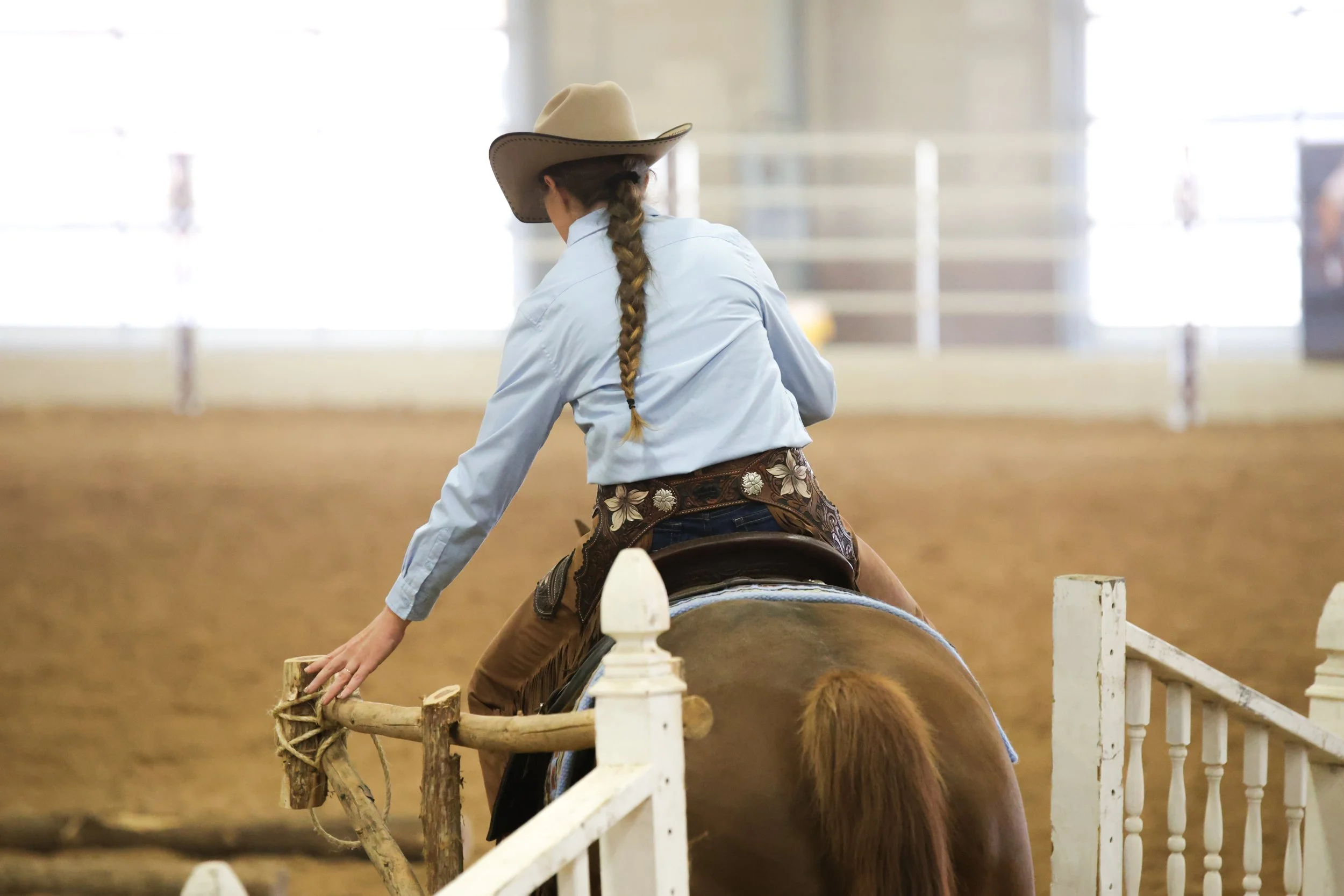A woman working the gate in a ranch trail course while riding a horse in an indoor arena, wearing a cowboy hat and western attire.