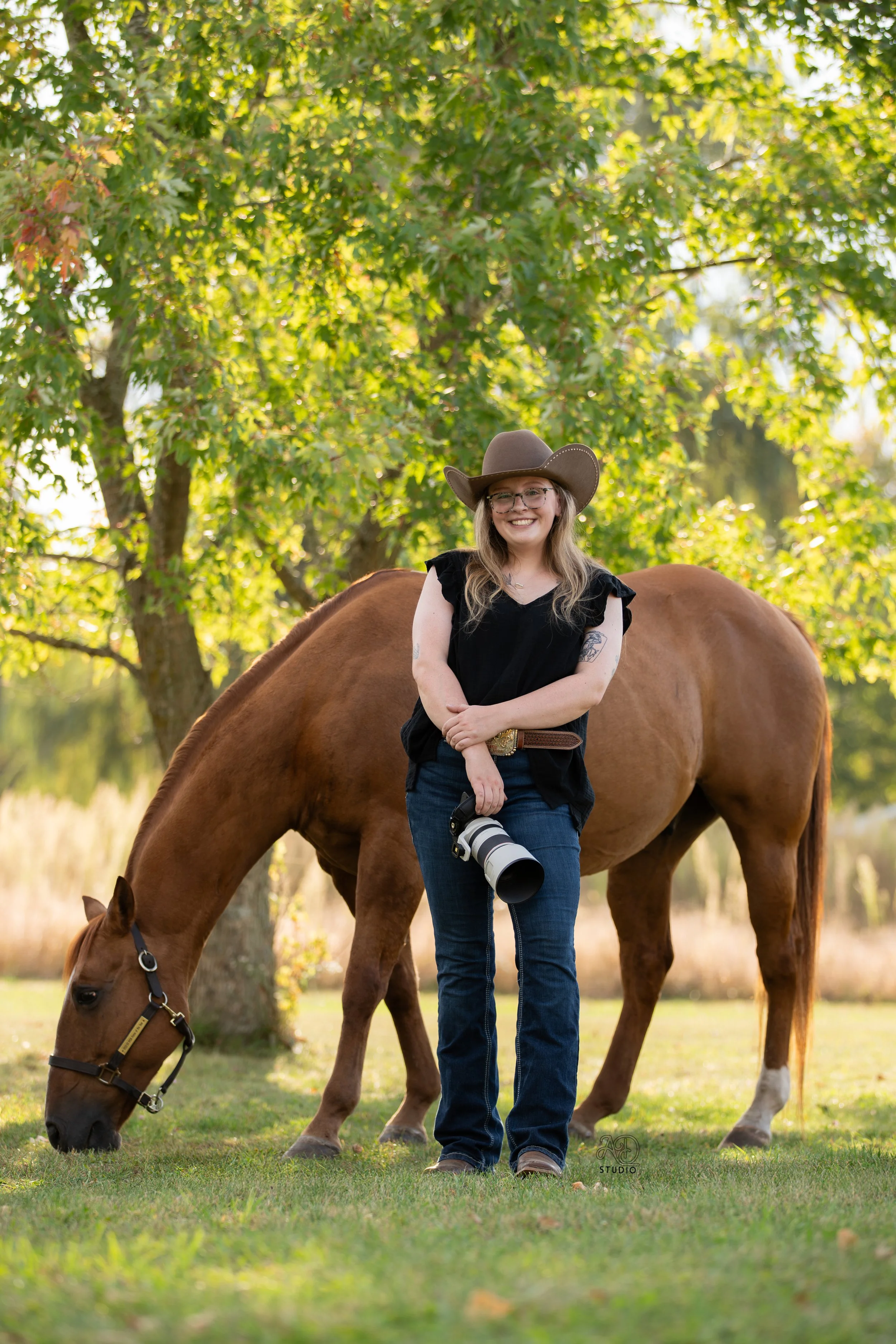 A woman with a camera stands outdoors next to a brown horse that is grazing. She is smiling, wearing a cowboy hat, glasses, a black top, and jeans, and is standing in front of a background of green trees and sunlight.